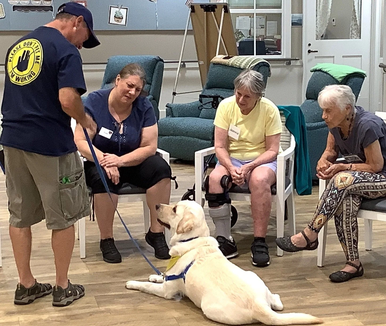 A service dog sits on the floor in front of four women seated in chairs. One woman has a prosthetic leg. The women are paying attention to the dog and the handler, who is standing and holding the dog's leash.