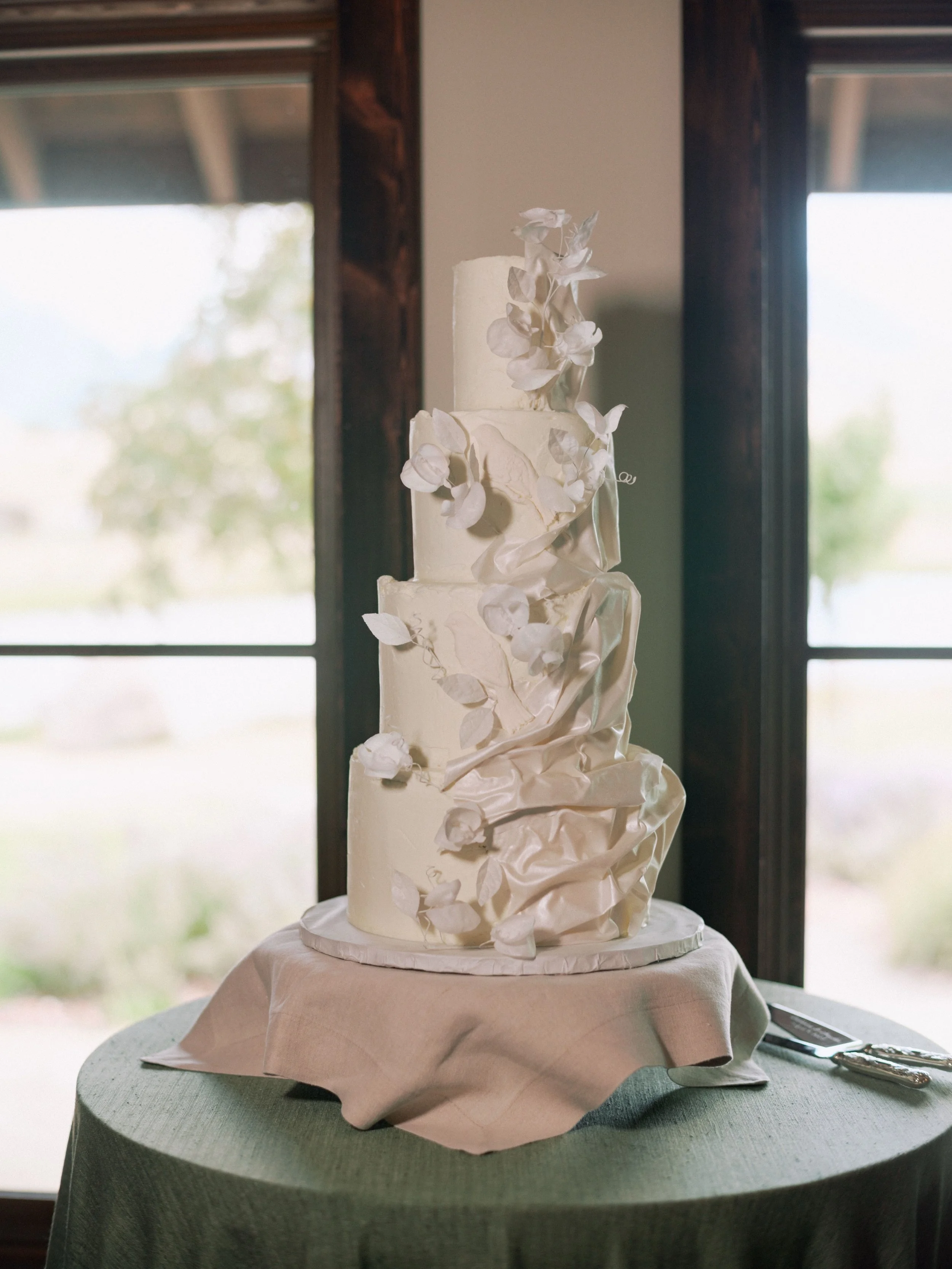 Cat + Hayden 🤍

Set against the beauty of Sage Lodge, this four-tiered cake was designed to feel soft, romantic, and full of movement. Edible silk drapery wraps each tier, with delicate modeling chocolate birds and hand-crafted wafer paper sweet pea