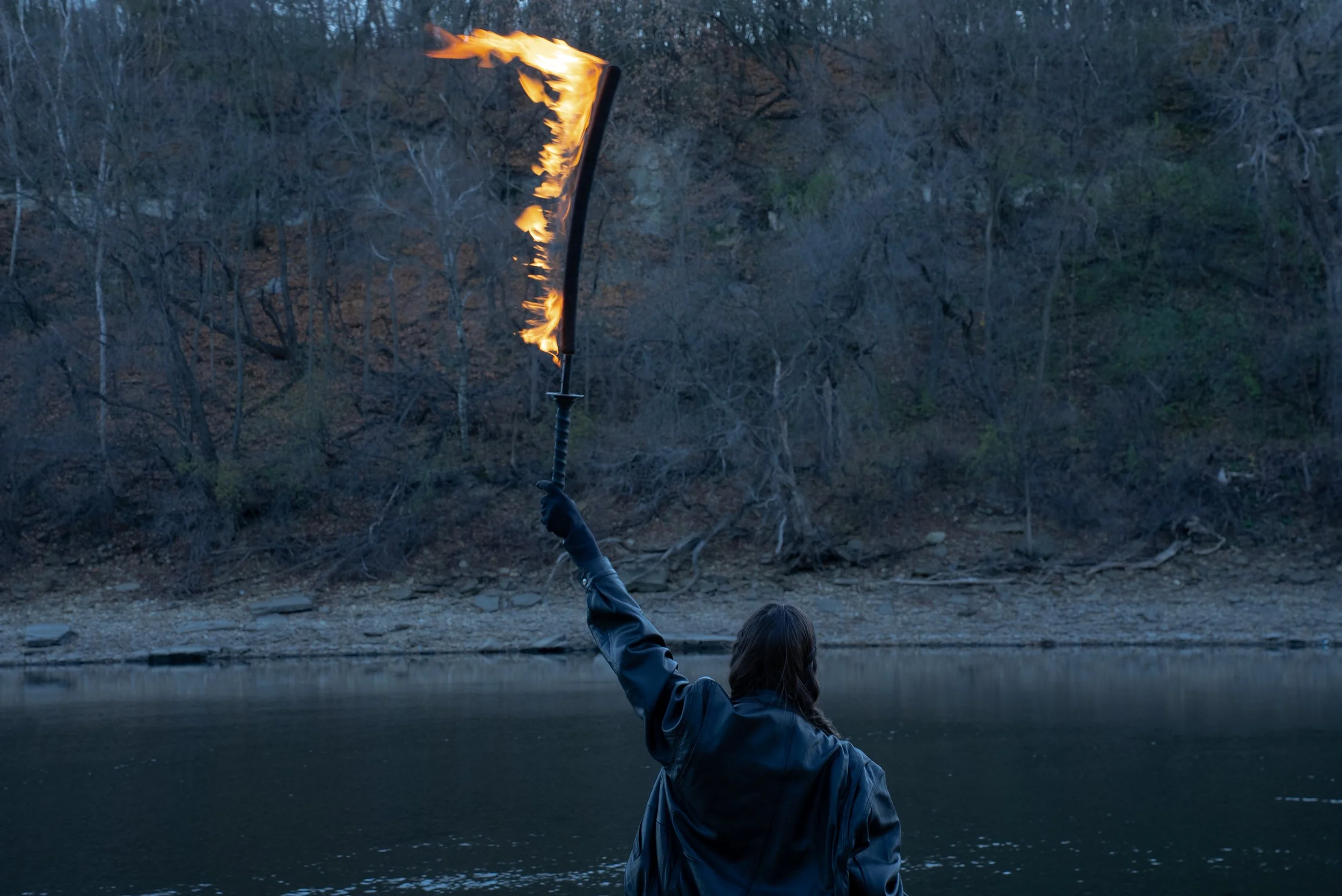 photo by Tori Schulte. An image of Nicole photogprahed from behind in an all black outfit, black leather jacket and black gloves with two braids holding a fire sword in the air in a victorious gesture at dusk near a valley in front of a river