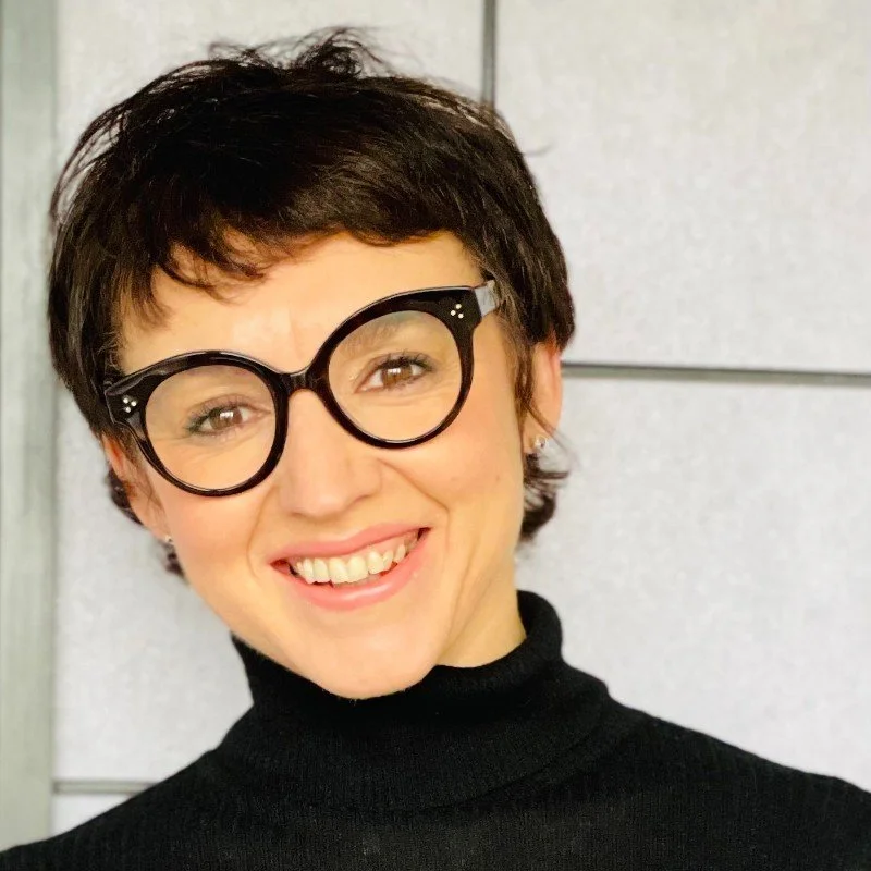 Woman with short dark hair wearing large round black glasses and a black turtleneck, smiling against a tiled wall background.