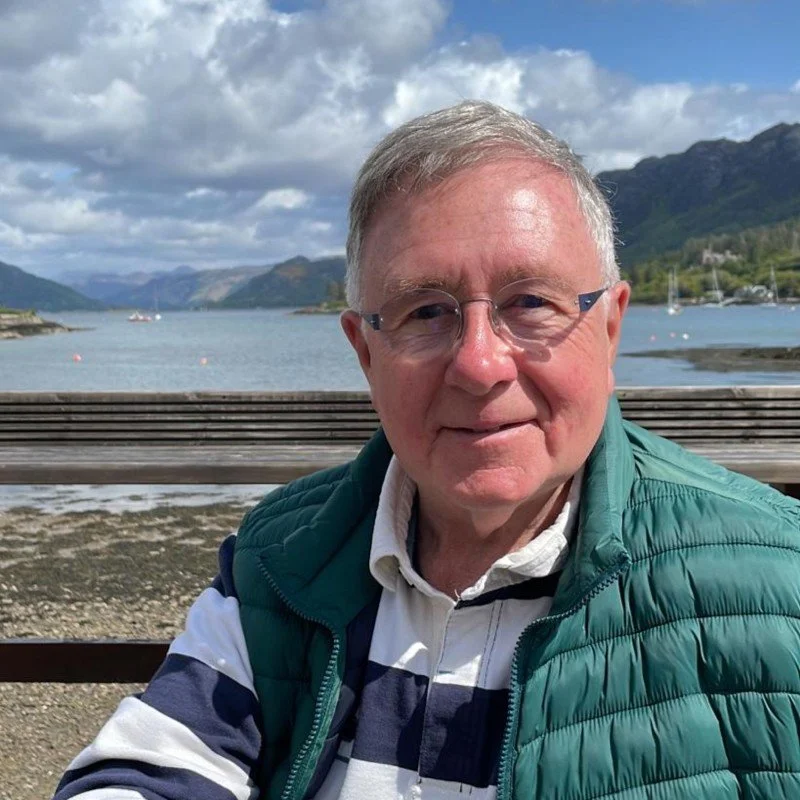 A man in glasses wearing a striped shirt and a green vest, sitting on a wooden bench by a body of water with mountains in the background under a partly cloudy sky.