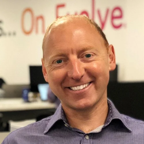 A smiling man with short blonde hair in a collared shirt, in an office setting.