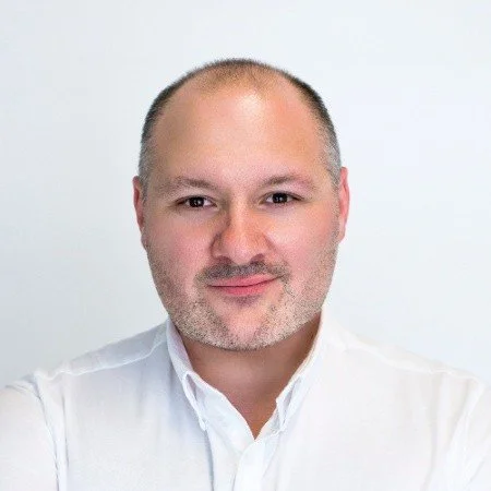 Close-up of a man with short hair and beard wearing a white shirt, smiling slightly against a plain white background.