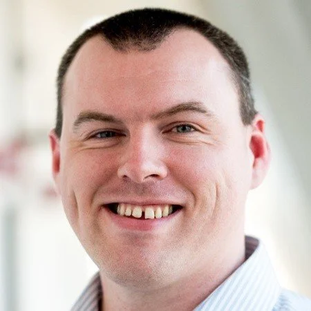 Close-up of a smiling man with short dark hair and a striped collared shirt.