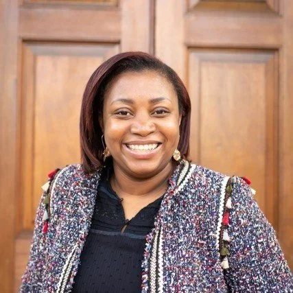 A woman with shoulder-length dark hair smiling, wearing a colorful, patterned jacket over a black top, standing in front of a wooden background.