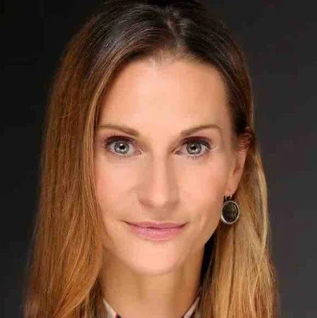 Close-up portrait of a woman with long, straight, light brown hair and earrings, smiling slightly against a dark background.