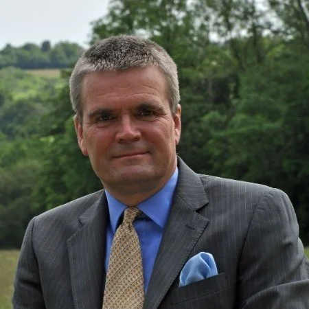 Portrait of a man in a gray suit and a blue shirt, outdoors with trees and greenery in the background.