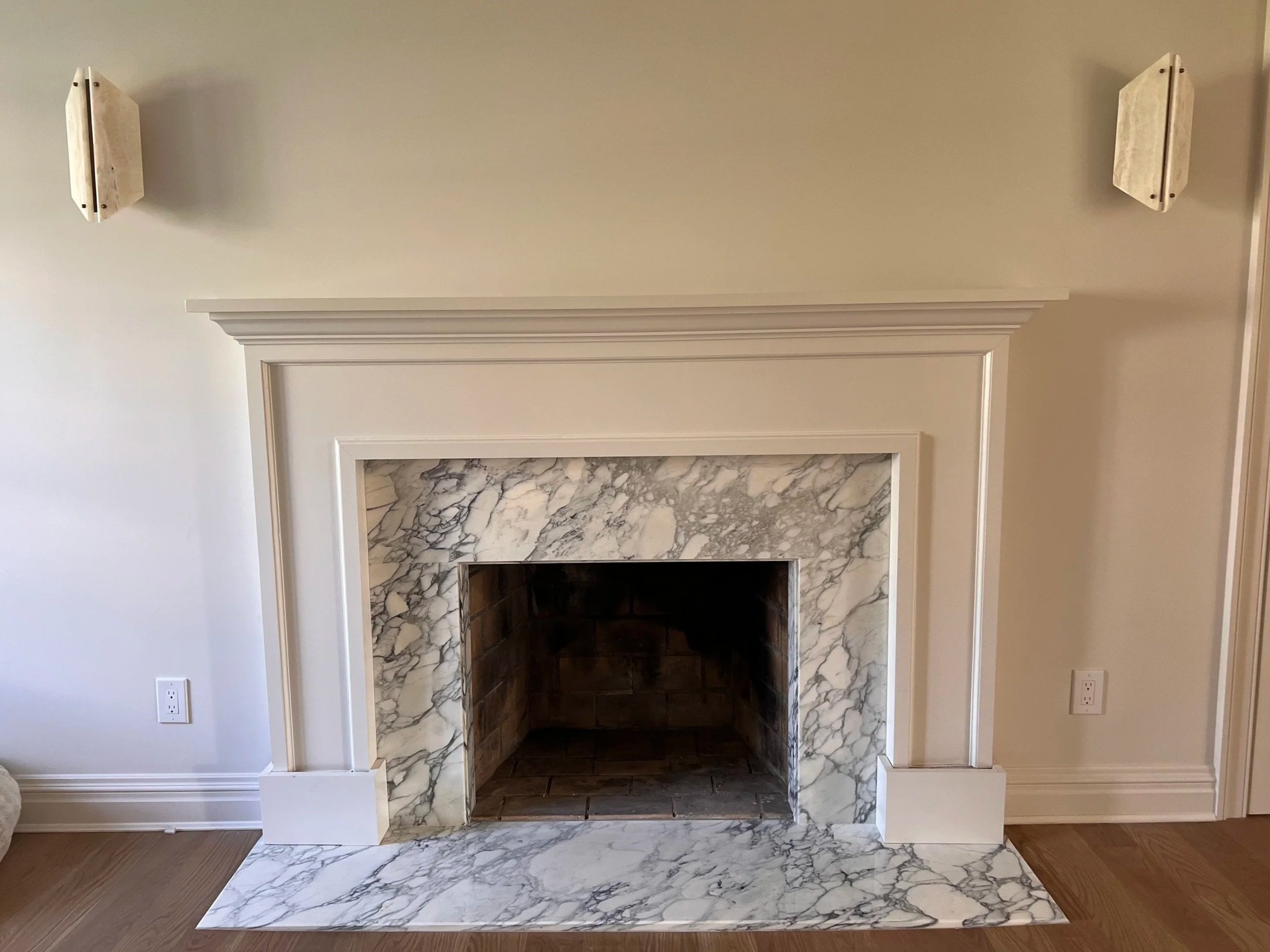 Empty fireplace with a marble surround and a mantel, set against a light-colored wall, with electrical outlets on either side and wood flooring.