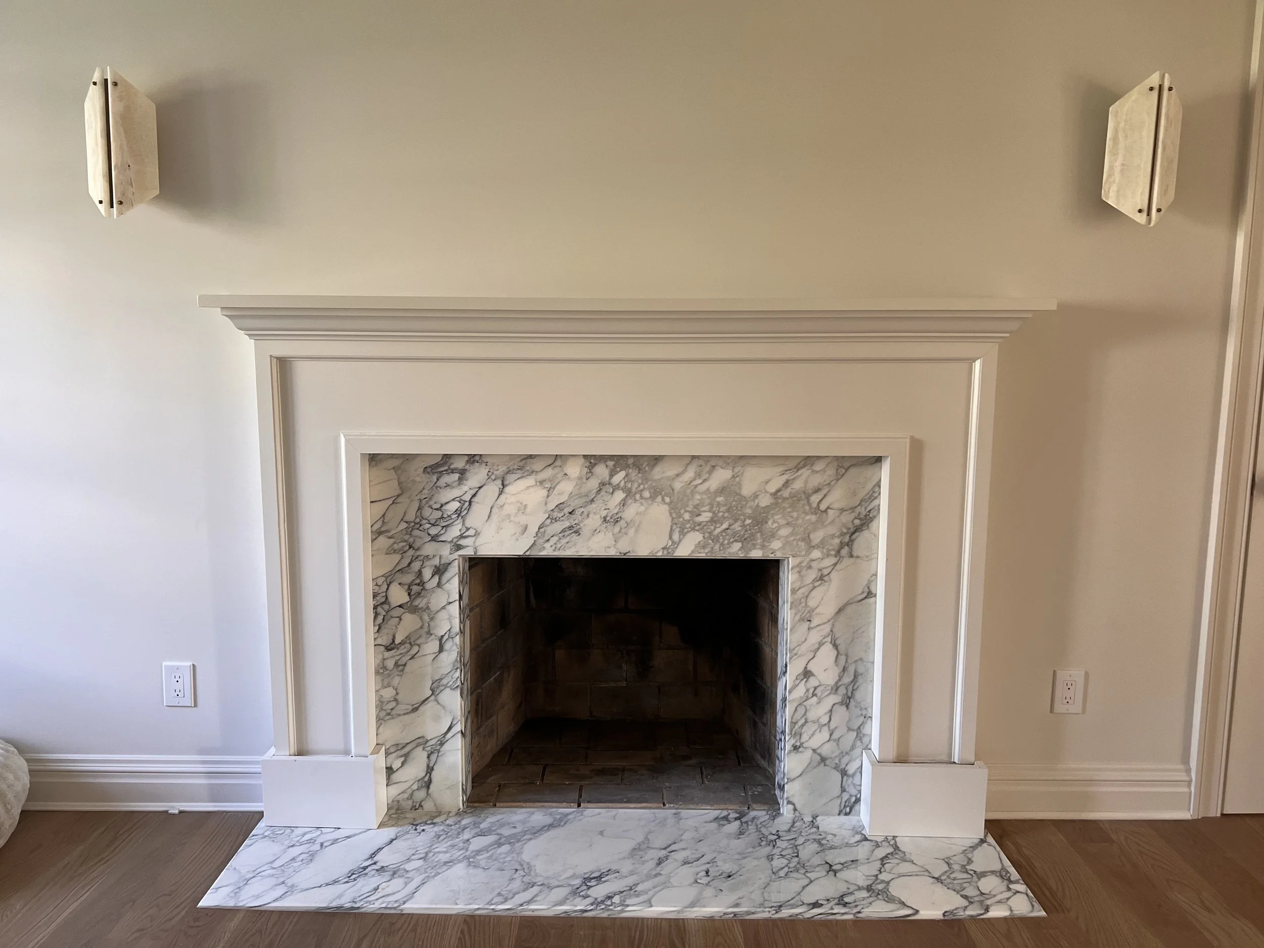 Fireplace with an Arabescato Turquoise marble surround and a mantel, set against a light-colored wall with two wall-mounted lights on either side in Port Washington, New York. 