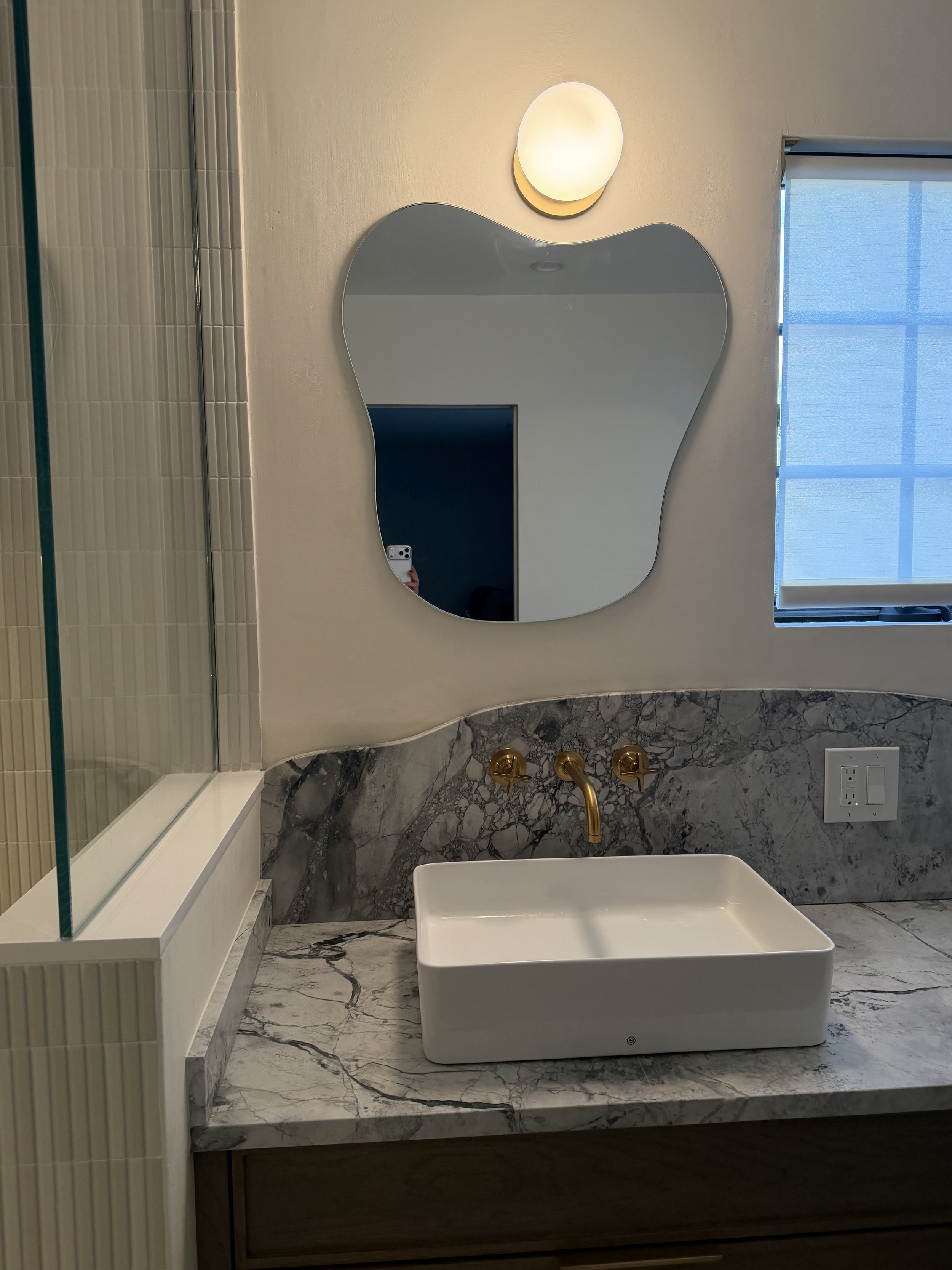 Modern bathroom in Roslyn, New York, with super white marble countertop, a large rectangular over-mount sink, a decorative mirror with an irregular shape, a wall-mounted light fixture, a window with blue blinds.