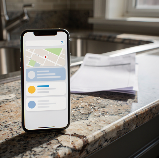 A smartphone displaying a map application sits on a kitchen counter next to a white mug, pen, and notebook.