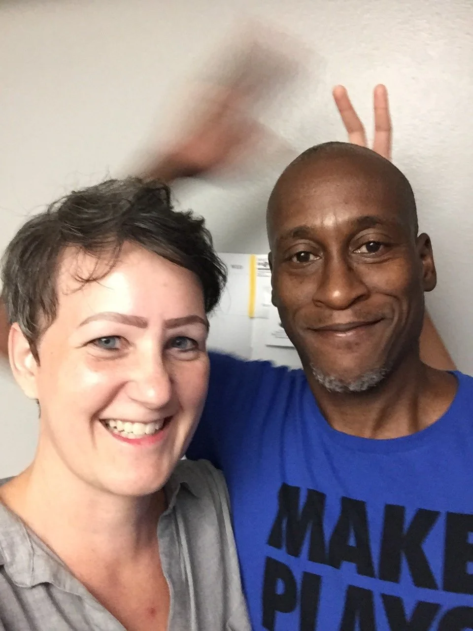 Smiling woman with short dark hair and a man with a shaved head and gray beard taking a selfie together in a room with a gray wall and some papers in the background. The woman's arm is raised with two fingers showing in a peace sign.