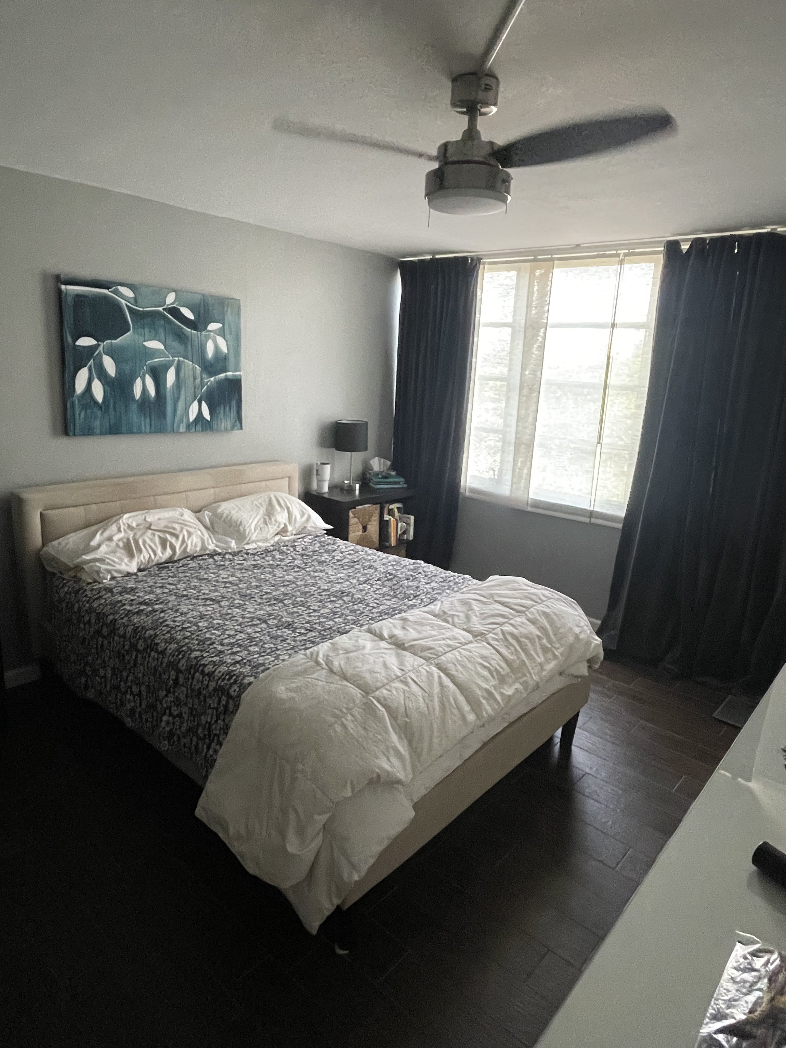 A bedroom with a beige headboard, white bedding, a dark patterned blanket, and an abstract blue and white painting above the bed. The room has dark curtains, a nightstand with books, a black lamp, and a window letting in natural light.
