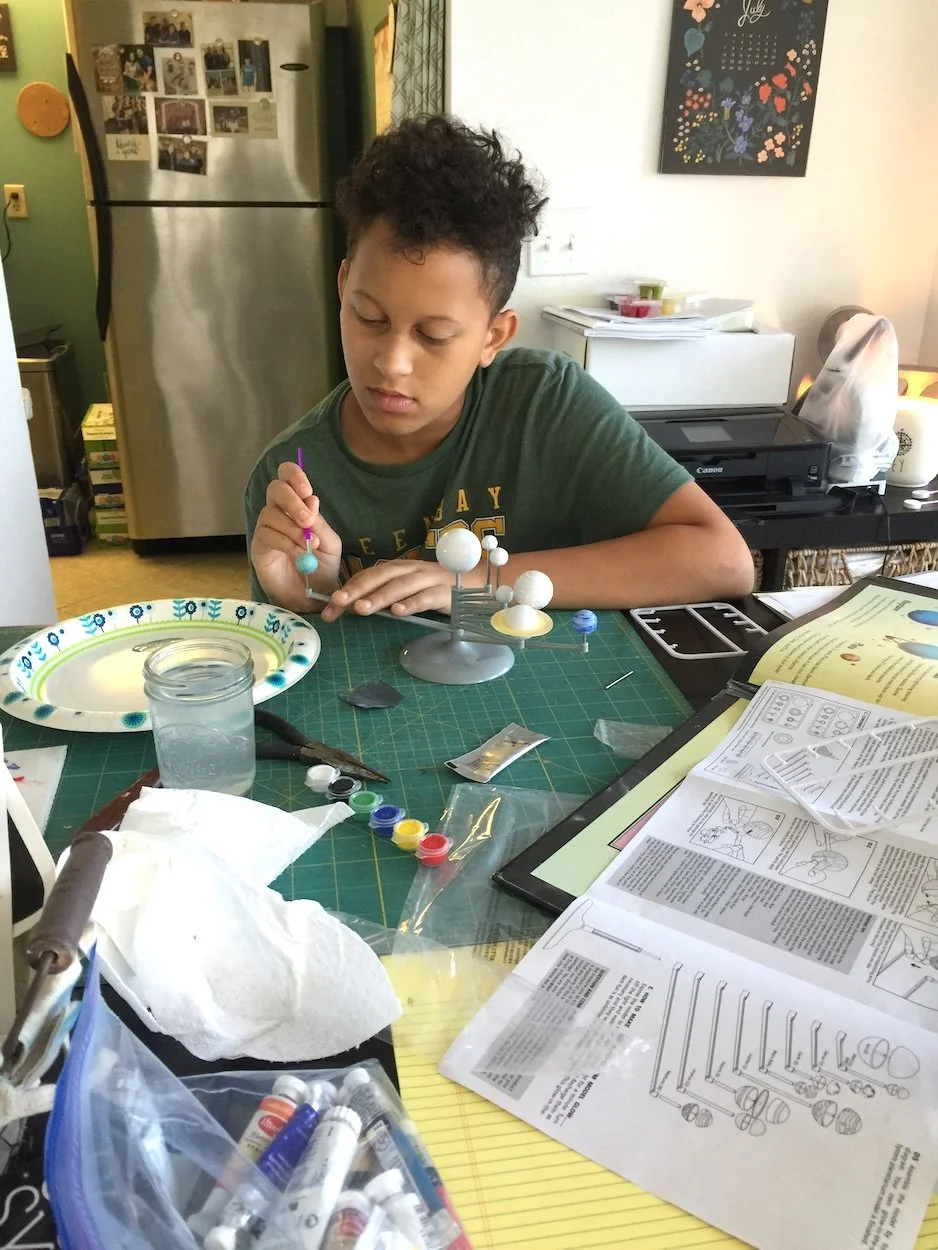 A boy sitting at a cluttered table working on a model solar system with paint and brushes, surrounded by modeling supplies and paperwork.