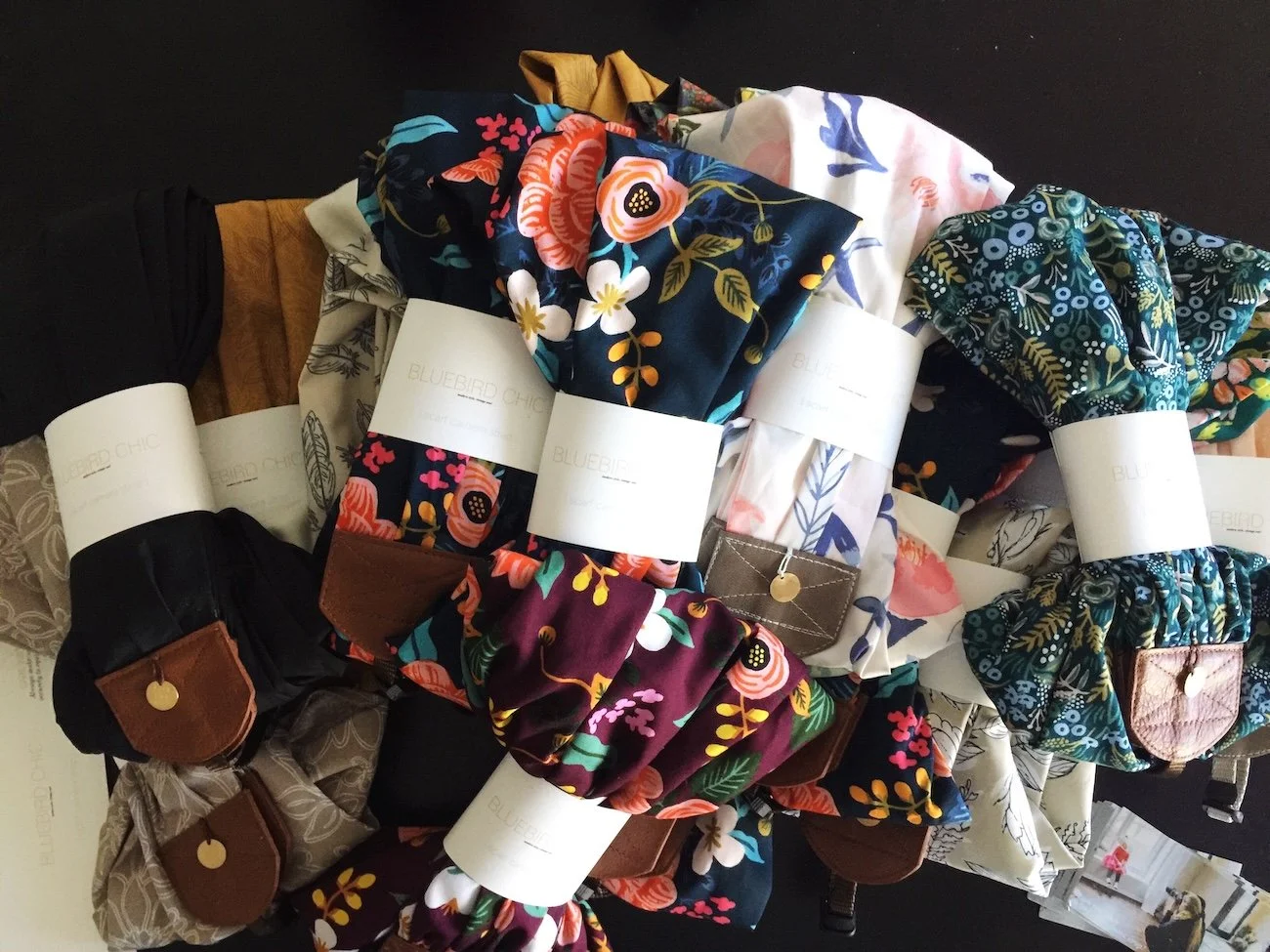 Collection of fabric scrunchies with floral and patterned designs, each with a white label wrapped around. Some scrunchies have leather or fabric accents.