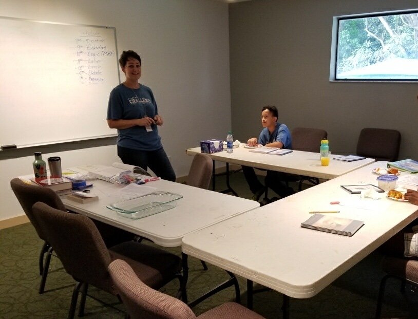 A woman standing near a whiteboard in a conference room, with a young boy sitting at a table. The room has a window showing trees outside and the table is cluttered with notebooks, water bottles, snacks, and a plastic container.