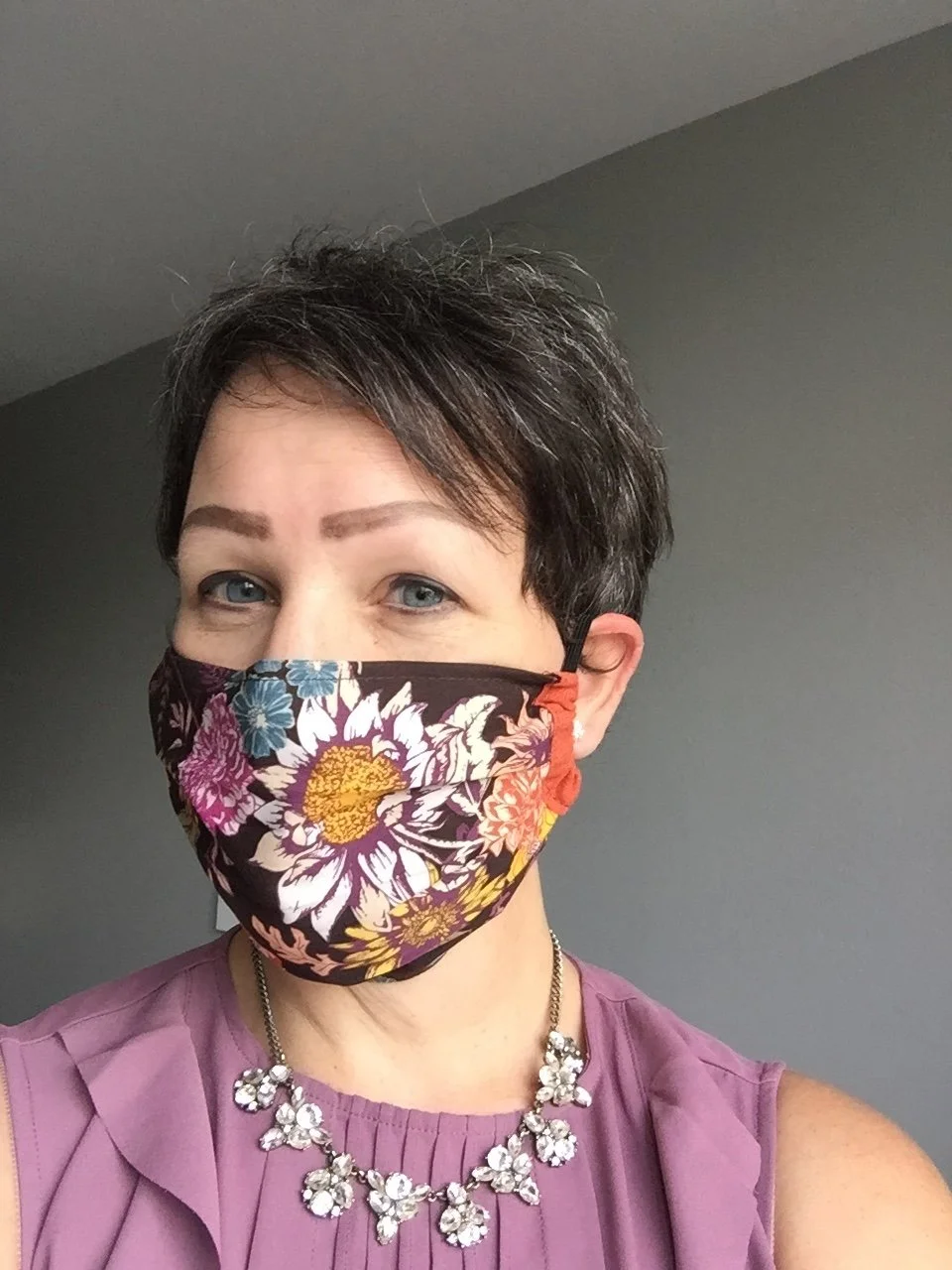 A woman with short dark hair and light blue eyes wearing a floral face mask, a purple sleeveless top, and a pearl and rhinestone necklace, standing indoors against a gray wall.