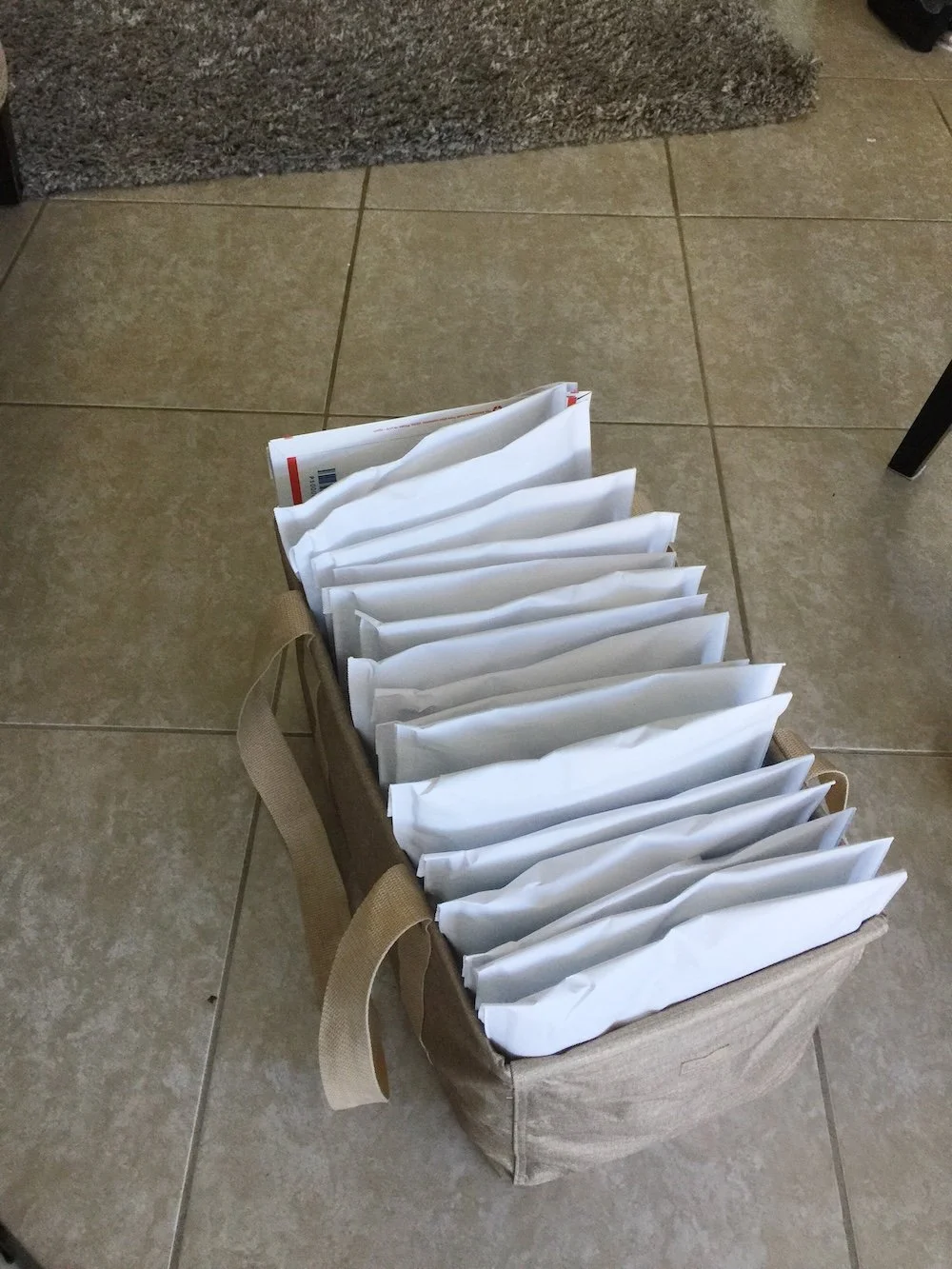 A reusable bag filled with multiple white envelopes or padded mailers, placed on a tiled floor.
