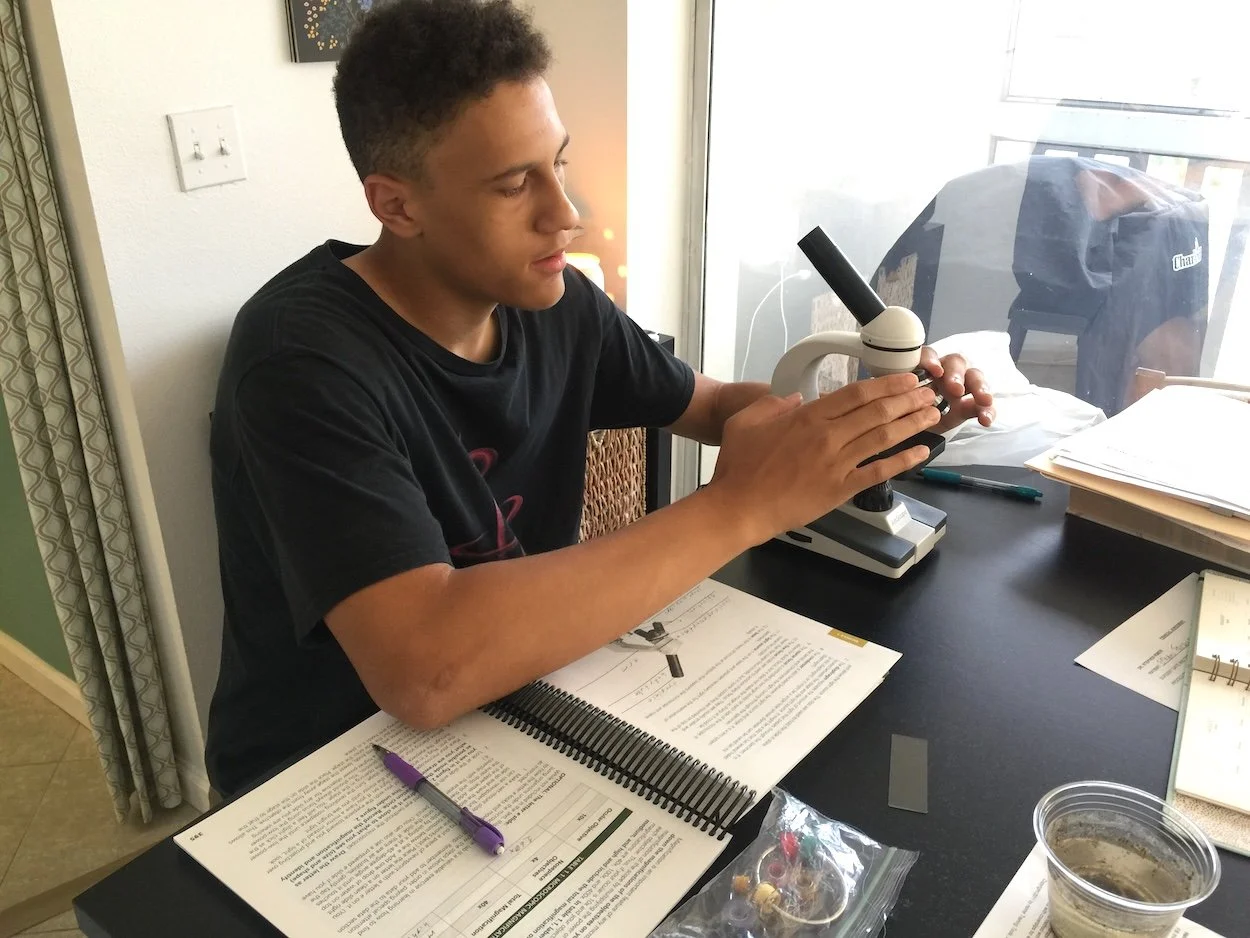 A young man sitting at a desk examining a slide under a microscope.