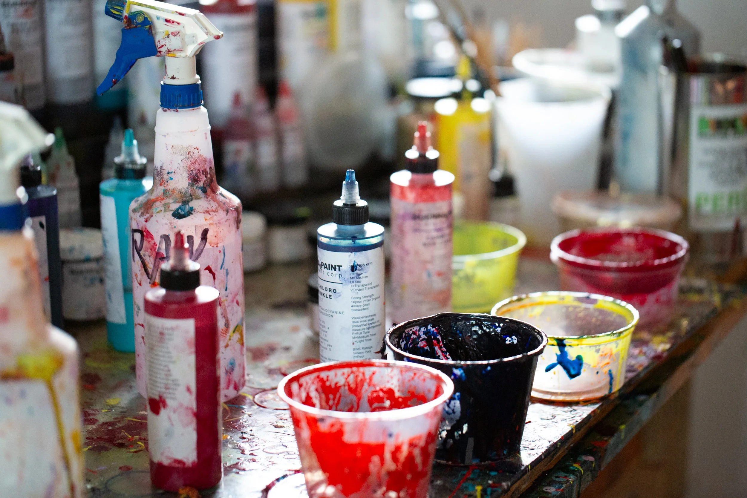 Close-up photo of an artist's work surface showing several paint bottles, spray bottles, and mixing cups filled with various colors including red, black, and yellow. The containers and surface are splattered with paint.