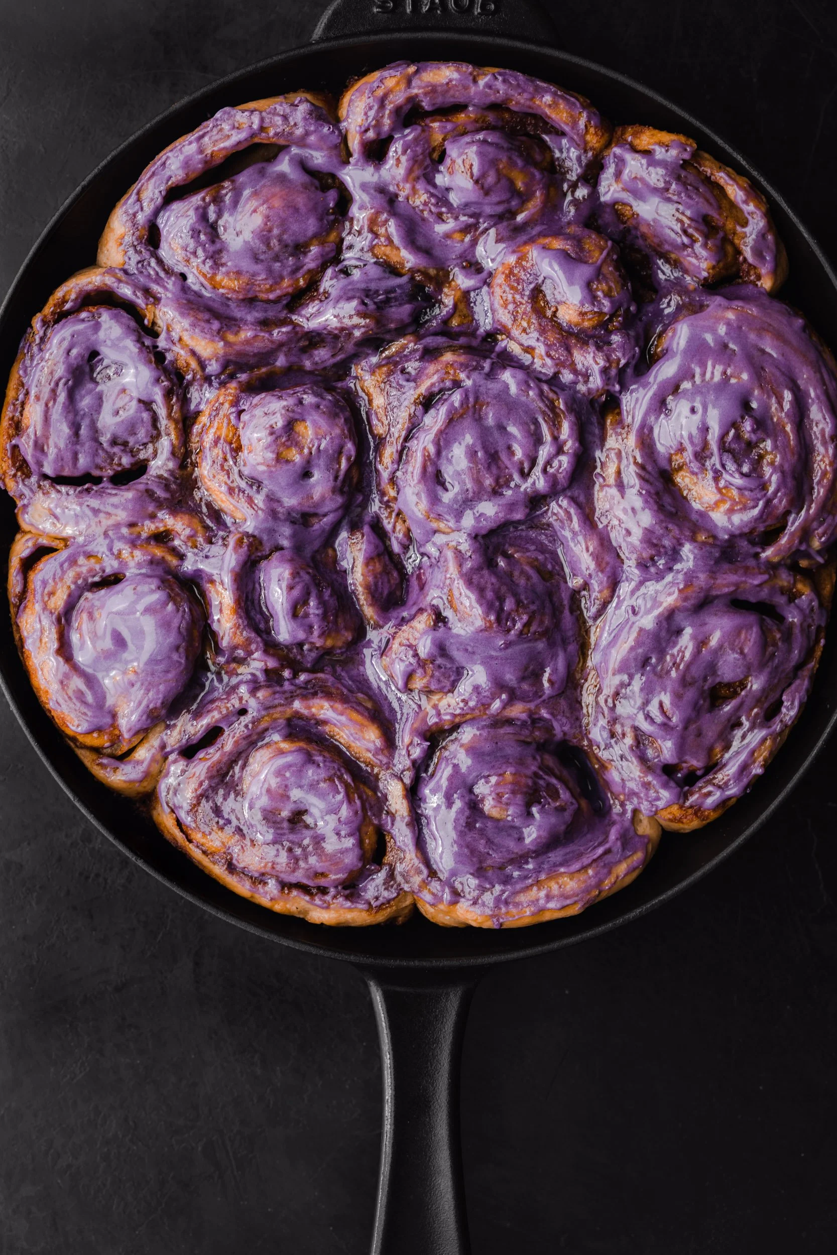 purple ube glazed cinnamon rolls in a black cast iron skillet on a black background 