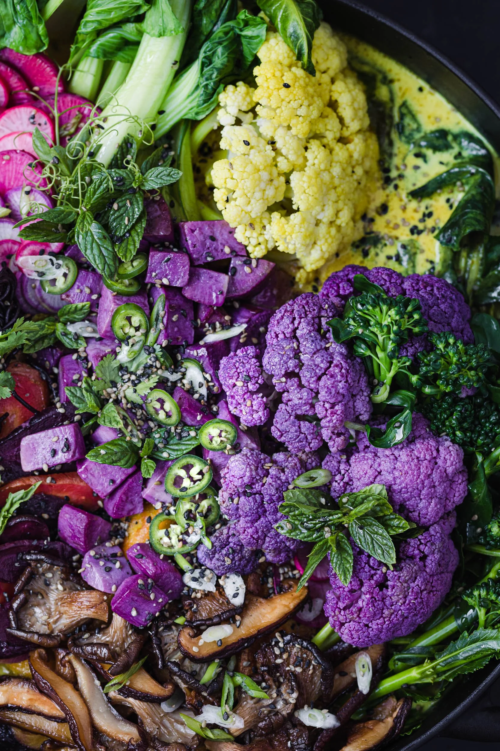 colorful veggie curry with purple cauliflower, greens, carrots, bok choy, radishes, mushrooms and more in a cast iron pot and grey background 
