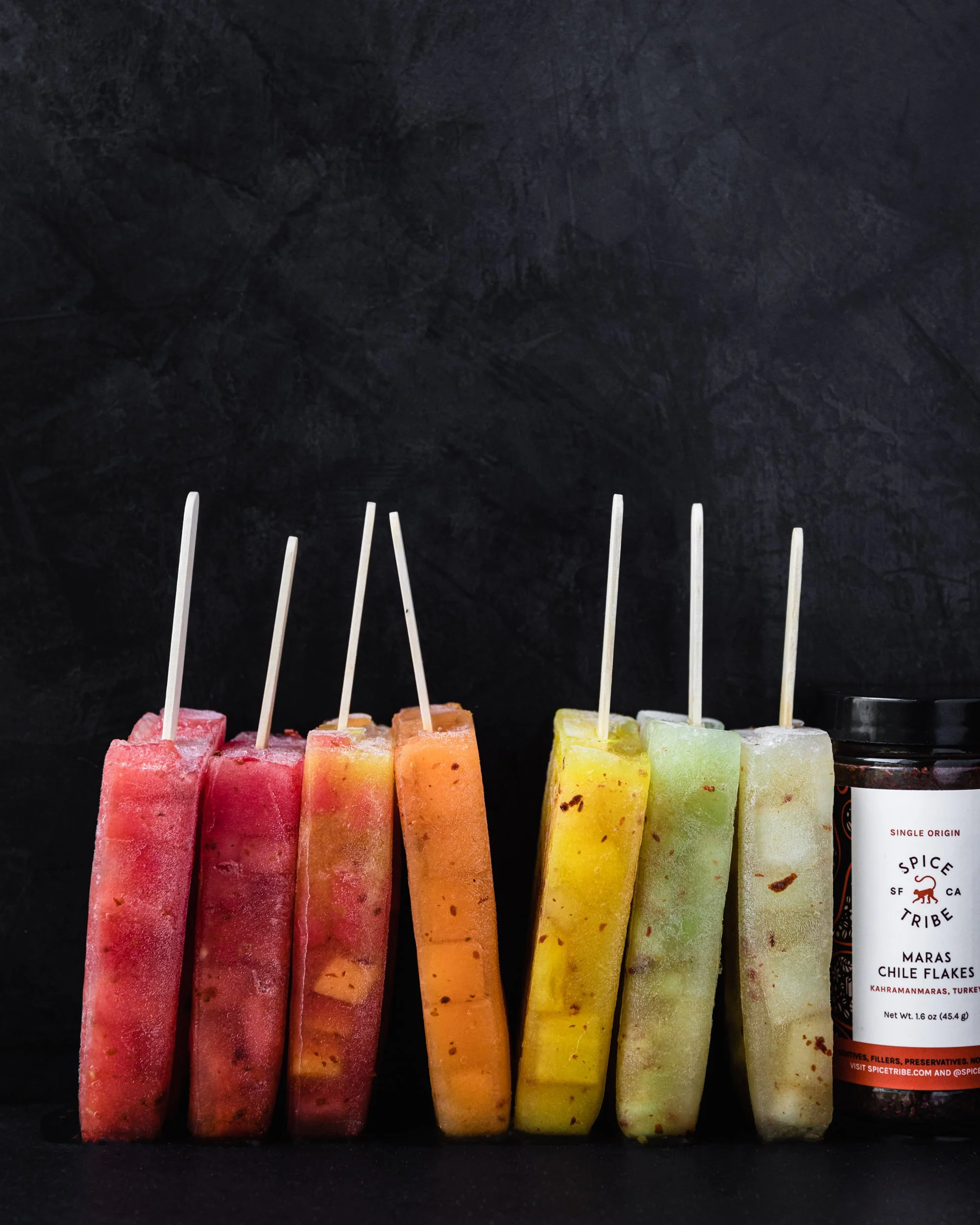 row of colorful melon popsicles with a chile flake bottle and black background 