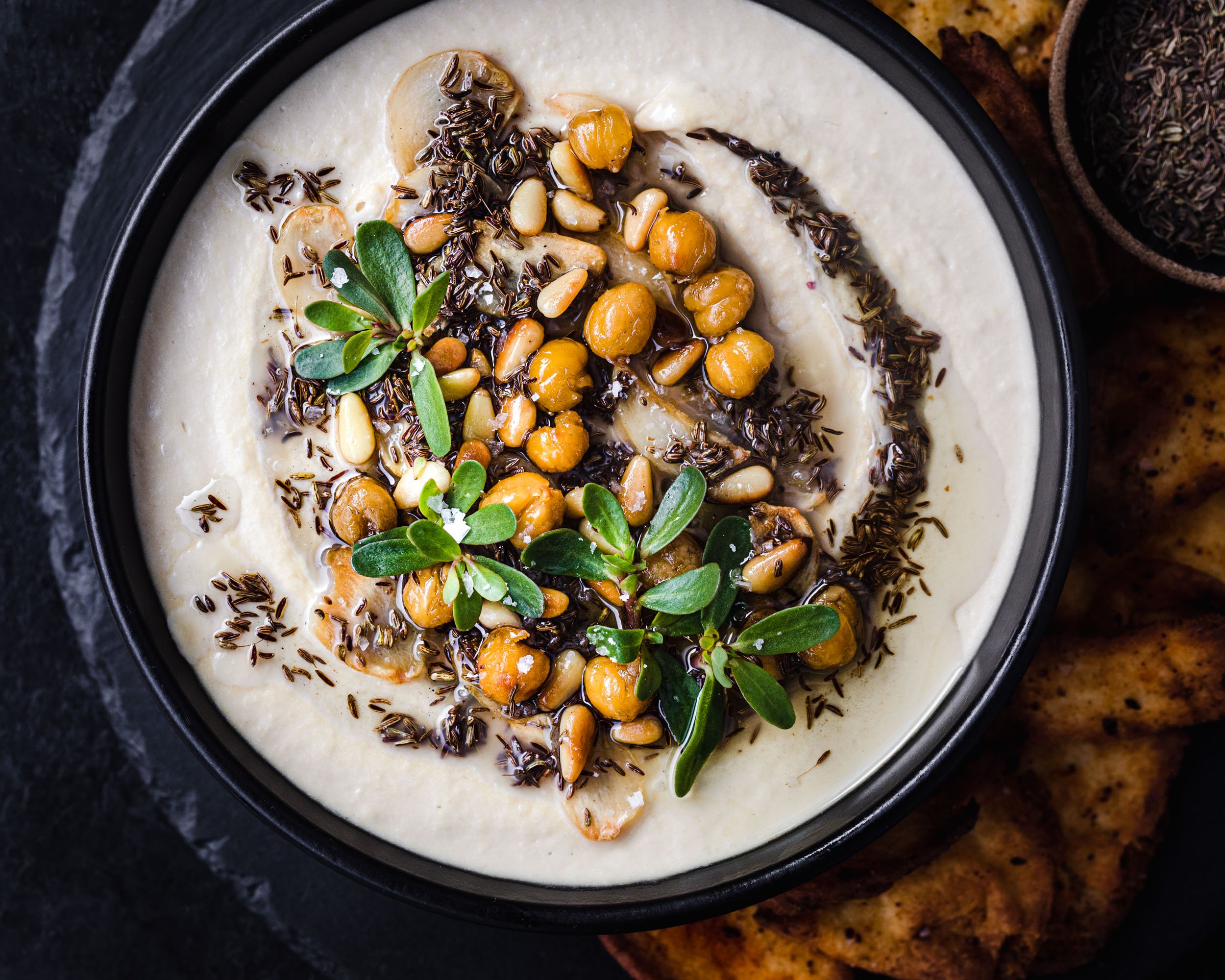 A bowl of hummus topped with olive, oil, cumin, herbs and flaky salt with pita chips on the side. 
