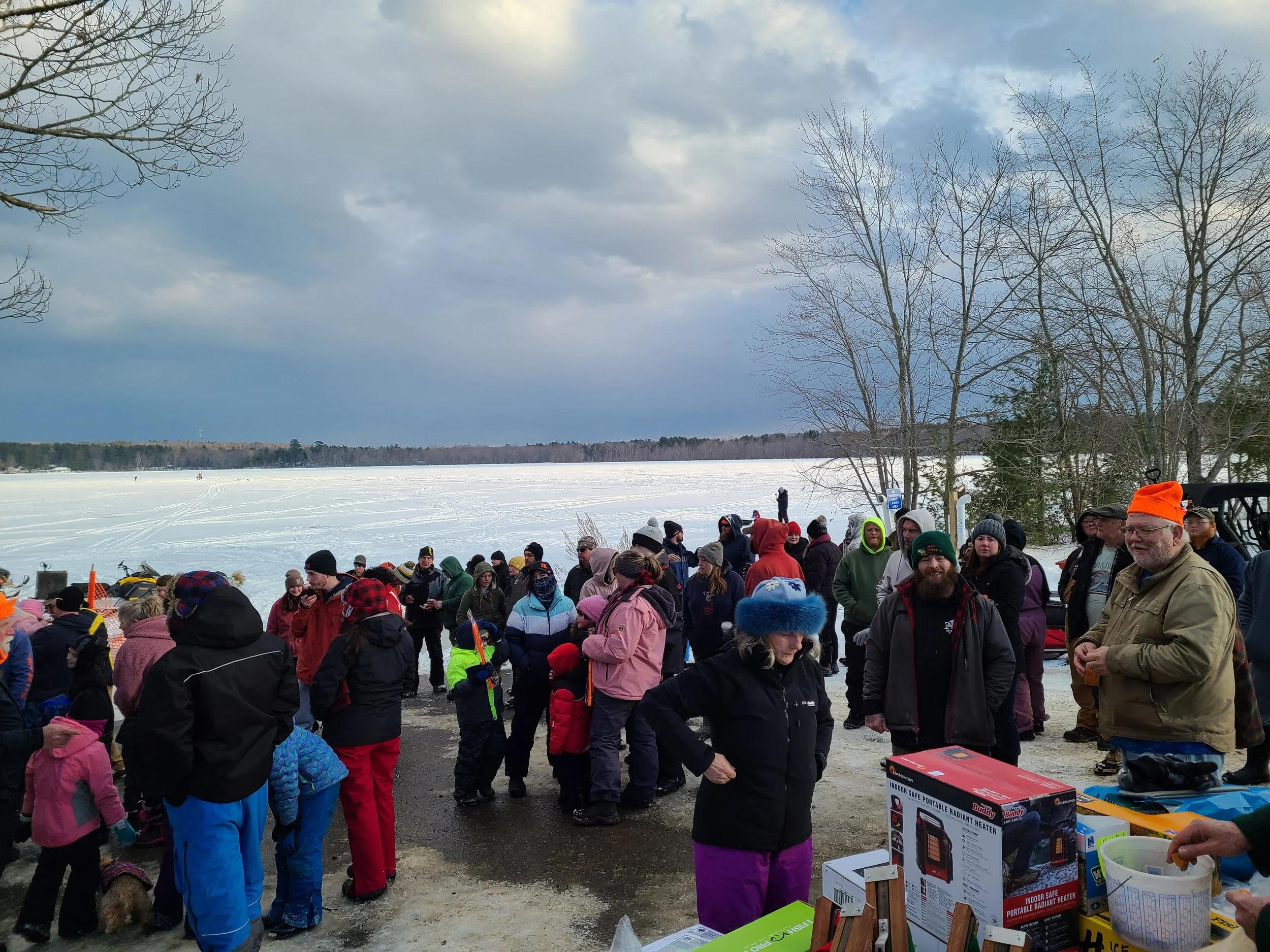 A group of people gathered outdoors on a winter day near a frozen lake with snow and trees, some wearing winter clothing and hats, some talking and standing in line, with boxes of miscellaneous items on a table.