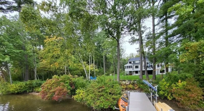 A lakeside scene with a dock, kayak, and paddleboard, surrounded by green trees and a white house in the background.