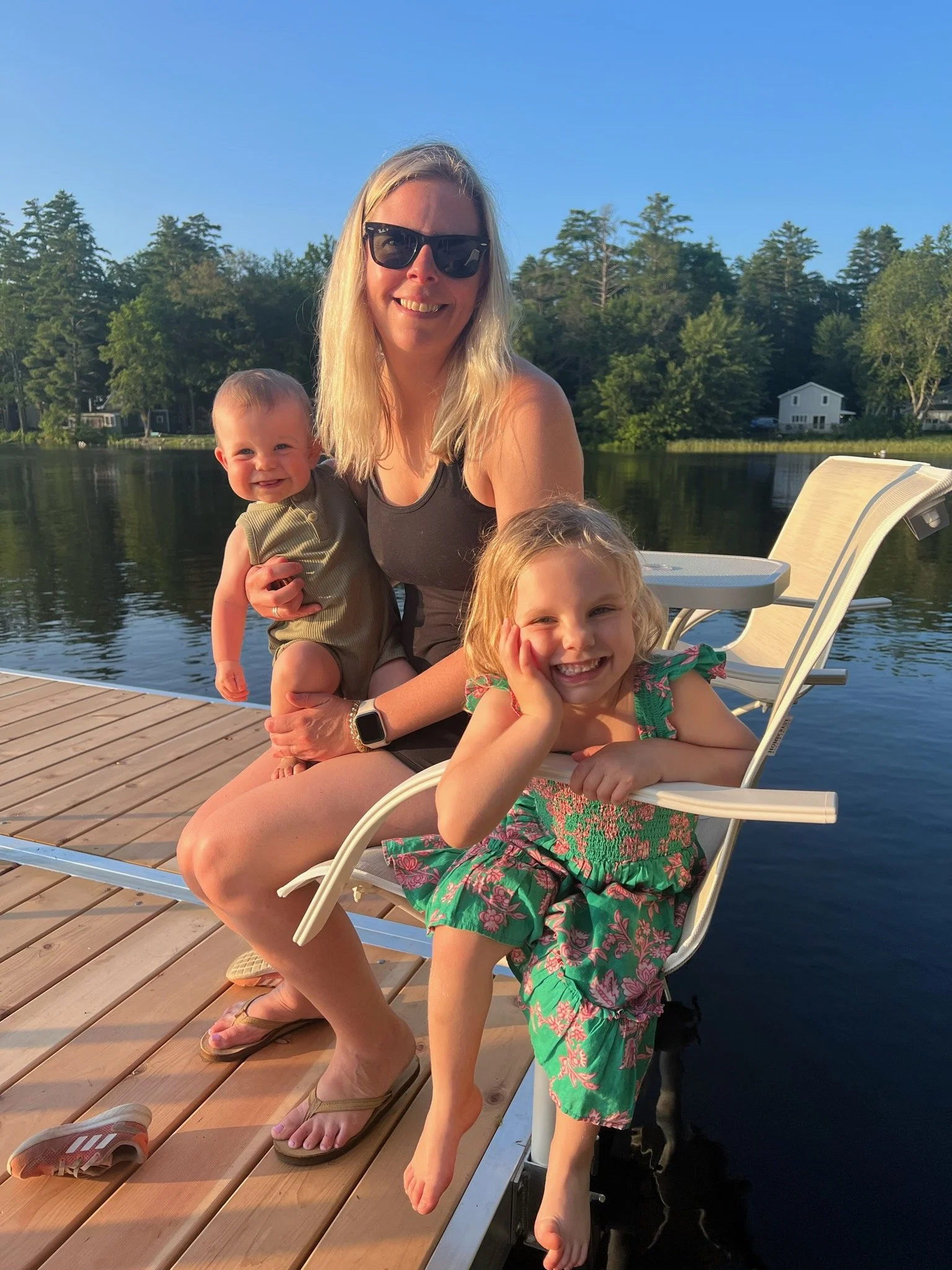 A woman with sunglasses and light hair sitting on a dock by the water, holding two young children, one on her lap and one sitting in a chair, all smiling on a sunny day.