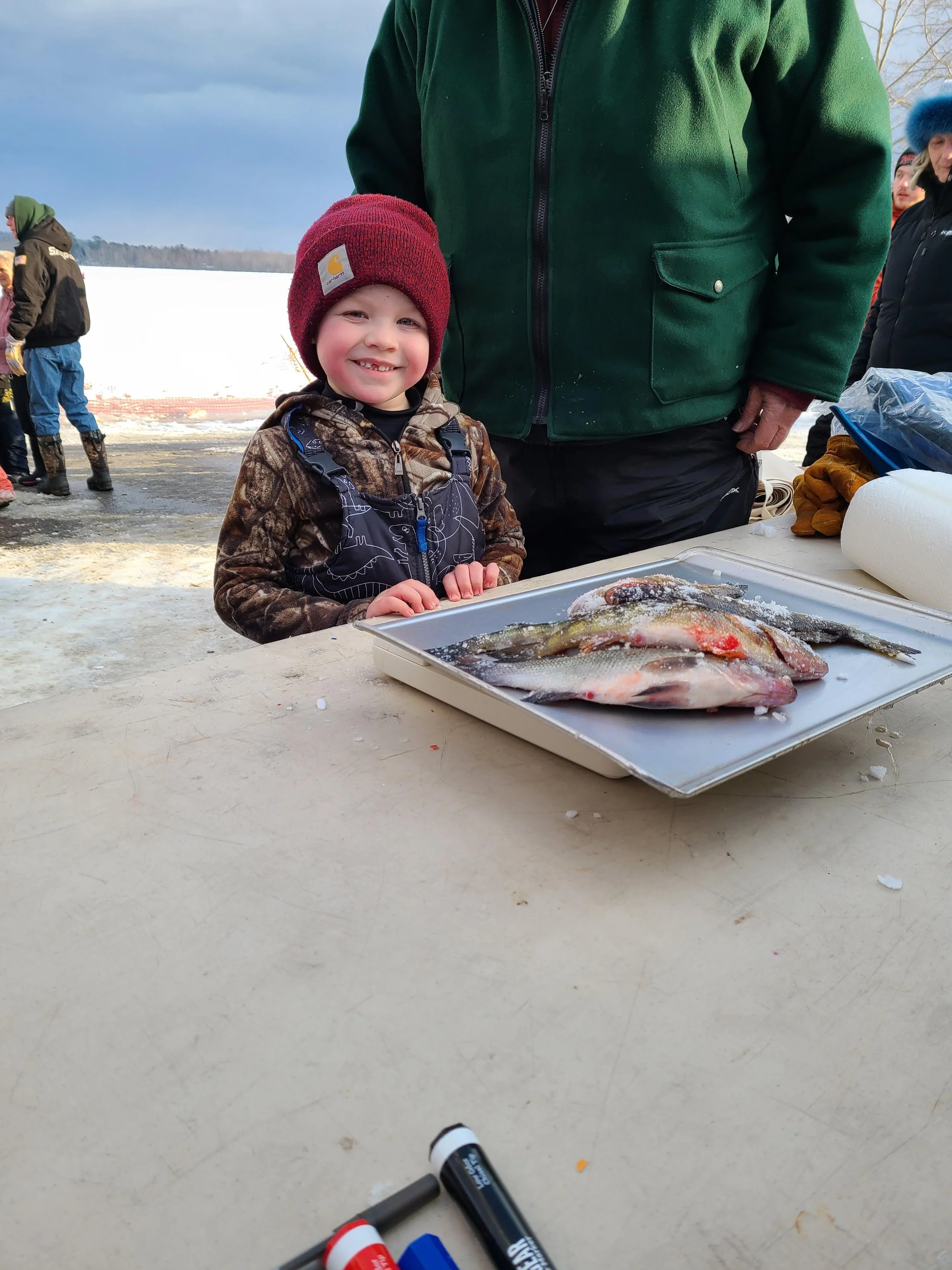 A young boy smiling and showing his teeth, wearing a red beanie and camouflage jacket, standing behind a table with freshly caught fish, outdoors on a snowy day, with an adult partially visible beside him. Other people are in the background.