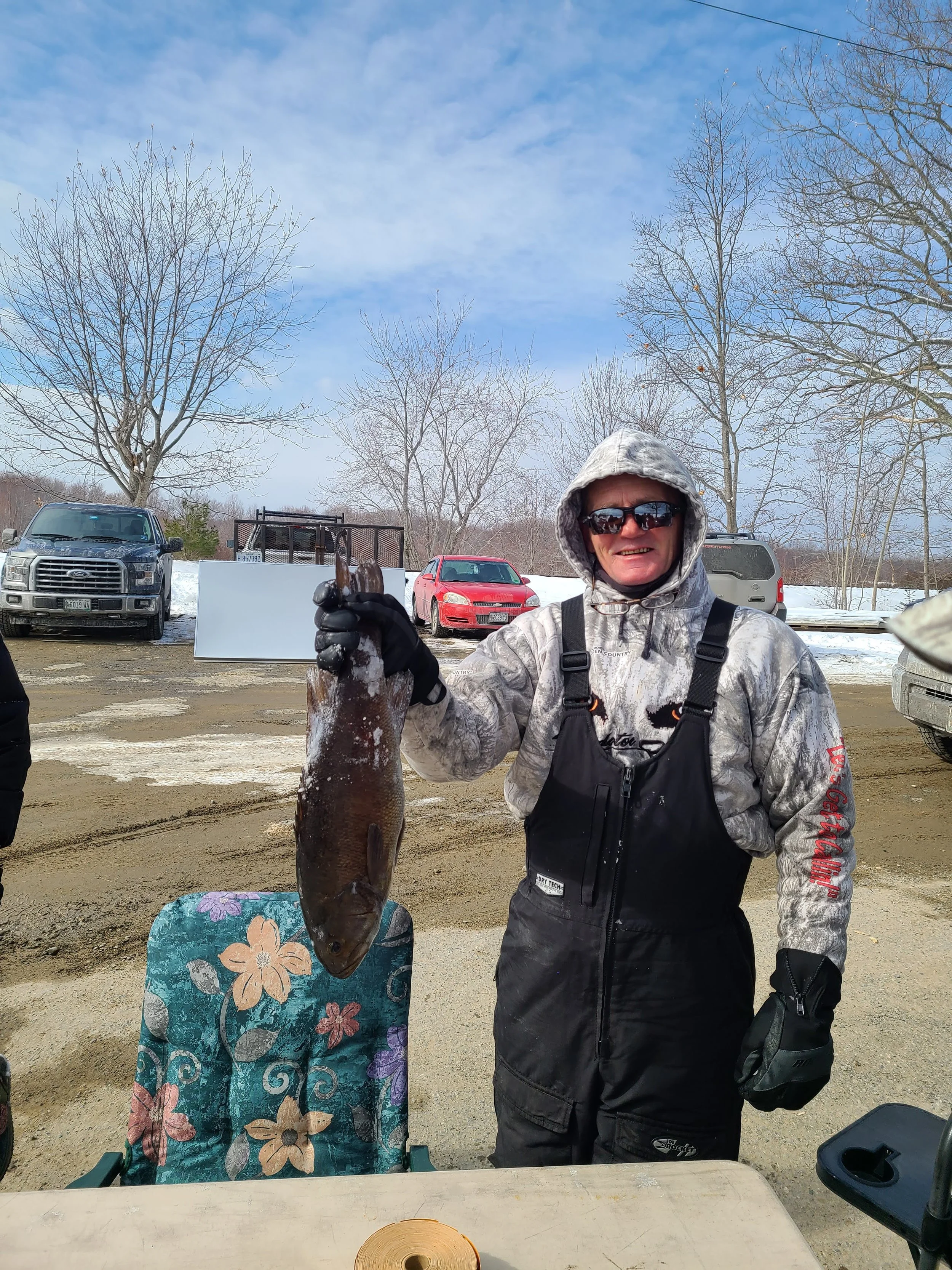 A man wearing winter gear and sunglasses holding a large fish outdoors in a parking lot with snow and leafless trees in the background.