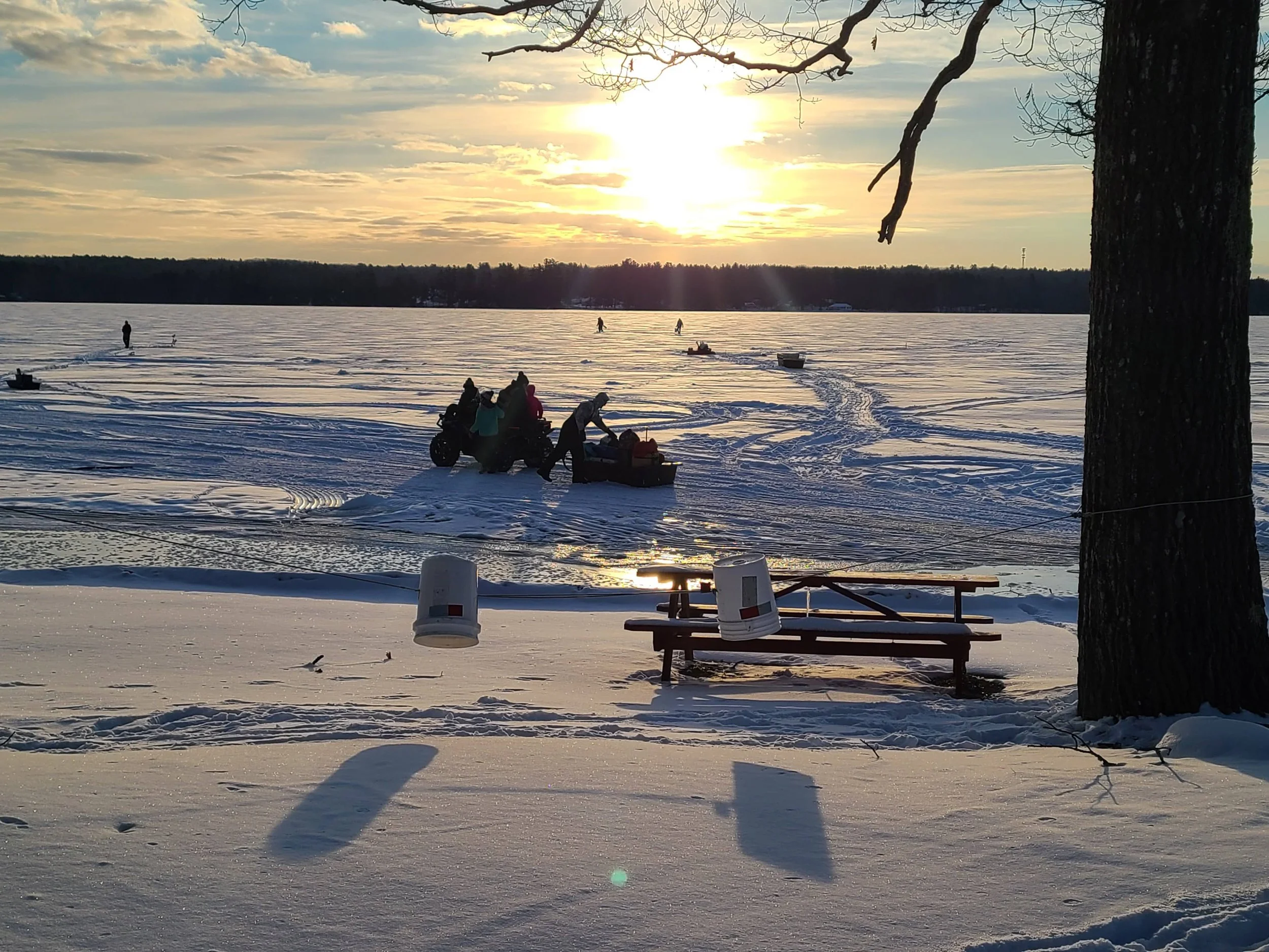 People riding snowmobiles on a Unity Pond during sunset, with snow-covered ground and leafless trees.