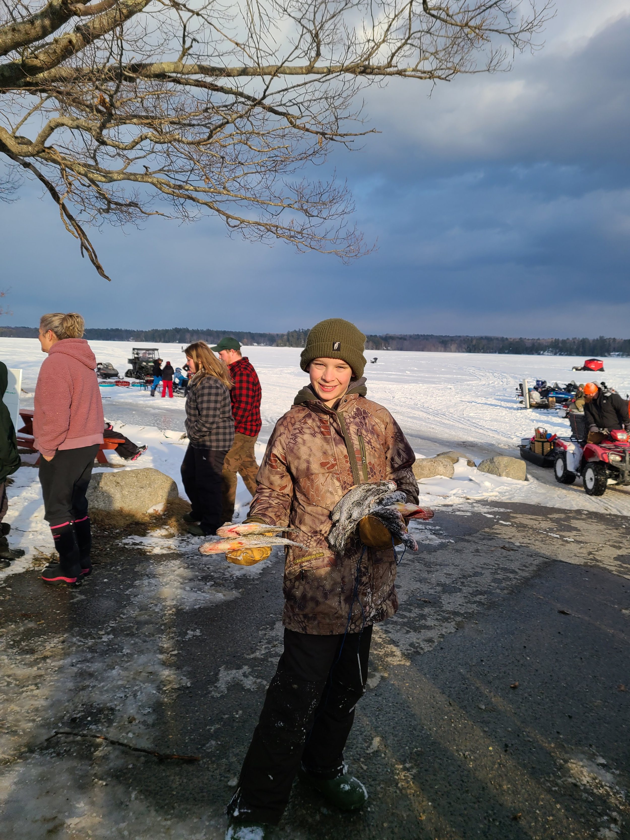 A boy holding a fish on a snowy lakeside with people and snowmobiles in the background.