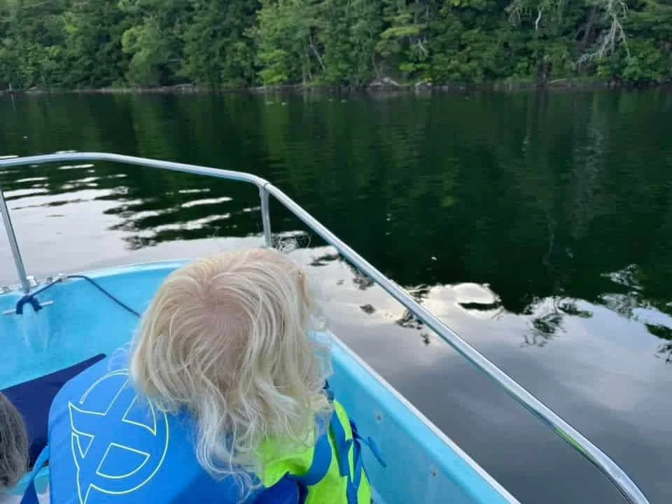 A person with curly blonde hair looking over the water on a boat, with trees and a shoreline in the distance.