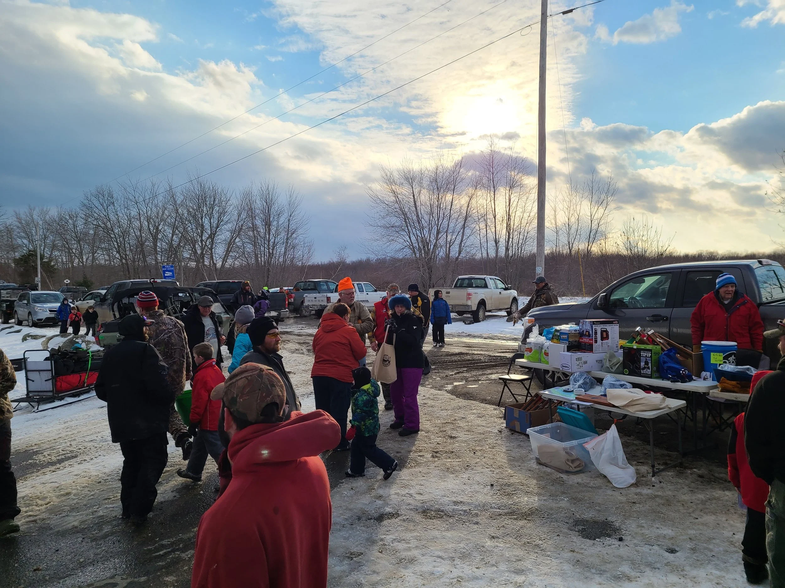 A group of people gathered outdoors in a parking lot, some wearing winter clothing, with tables set up with various supplies, and cars parked in the background under a cloudy sky with sunlight breaking through.