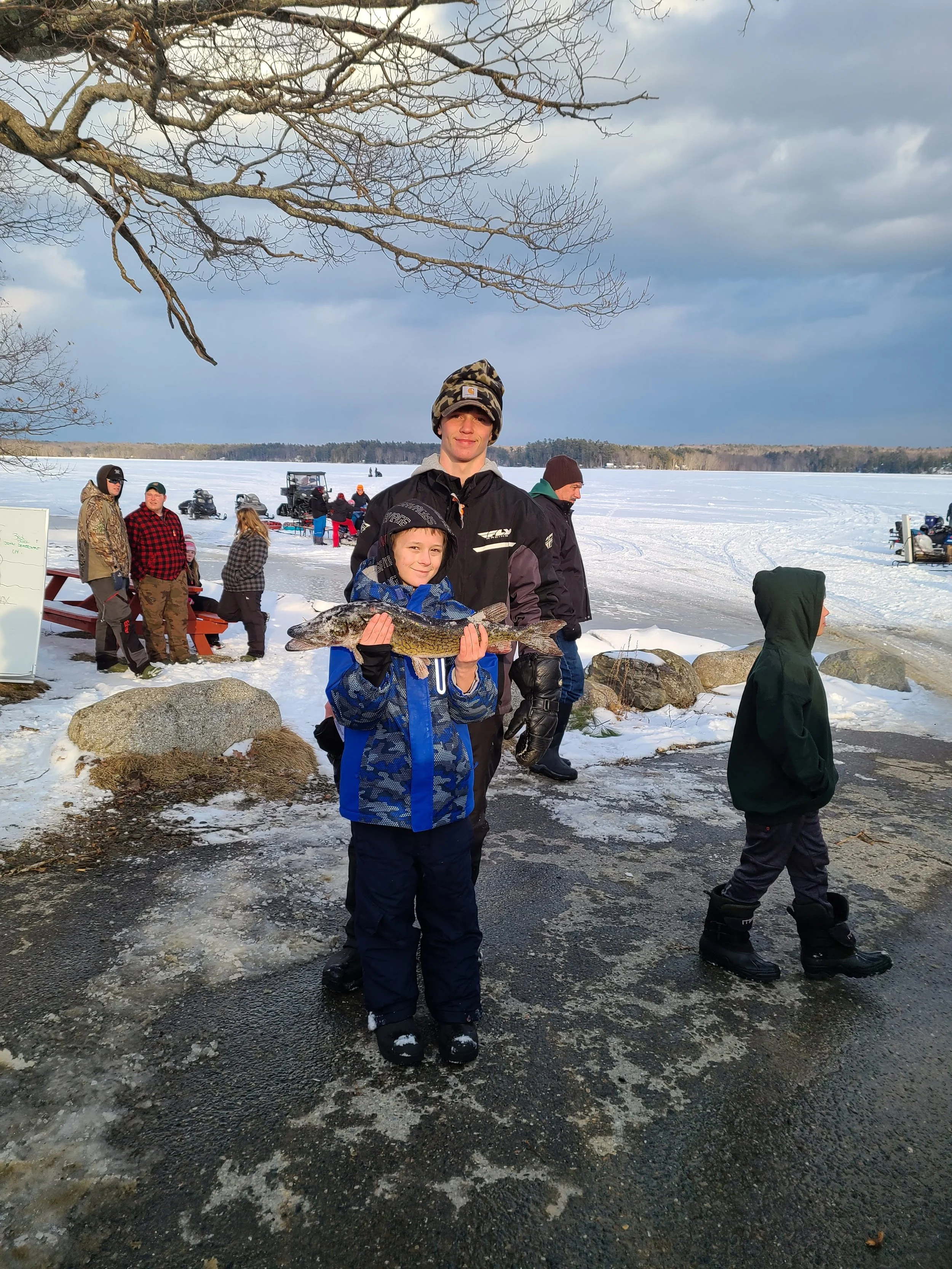 A boy holding a large fish with an older boy standing behind him, on a snowy lakeside with other people and snowmobiles in the background.