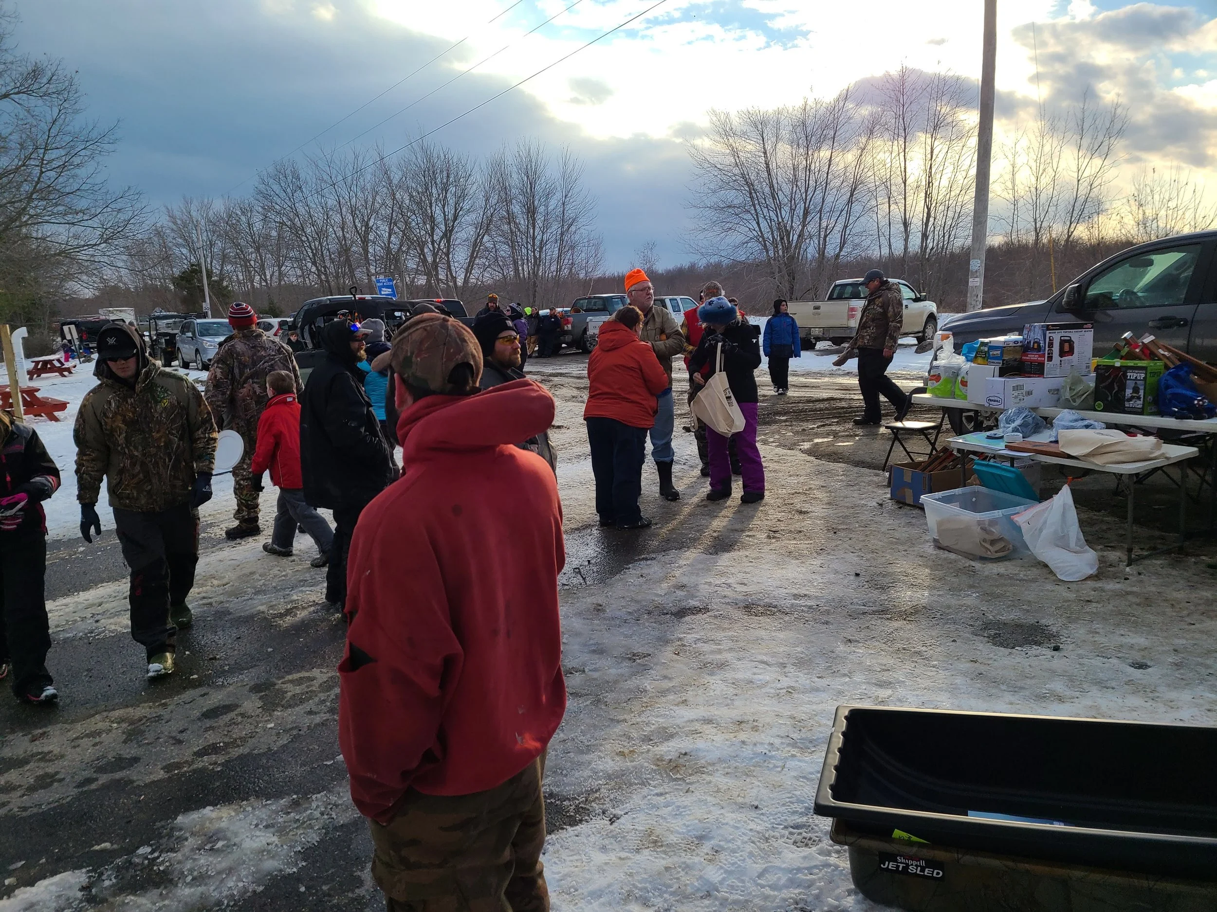 People gathered outdoors on a snowy day, some browsing tables with items for sale or giveaway, with parked cars and leafless trees in the background.