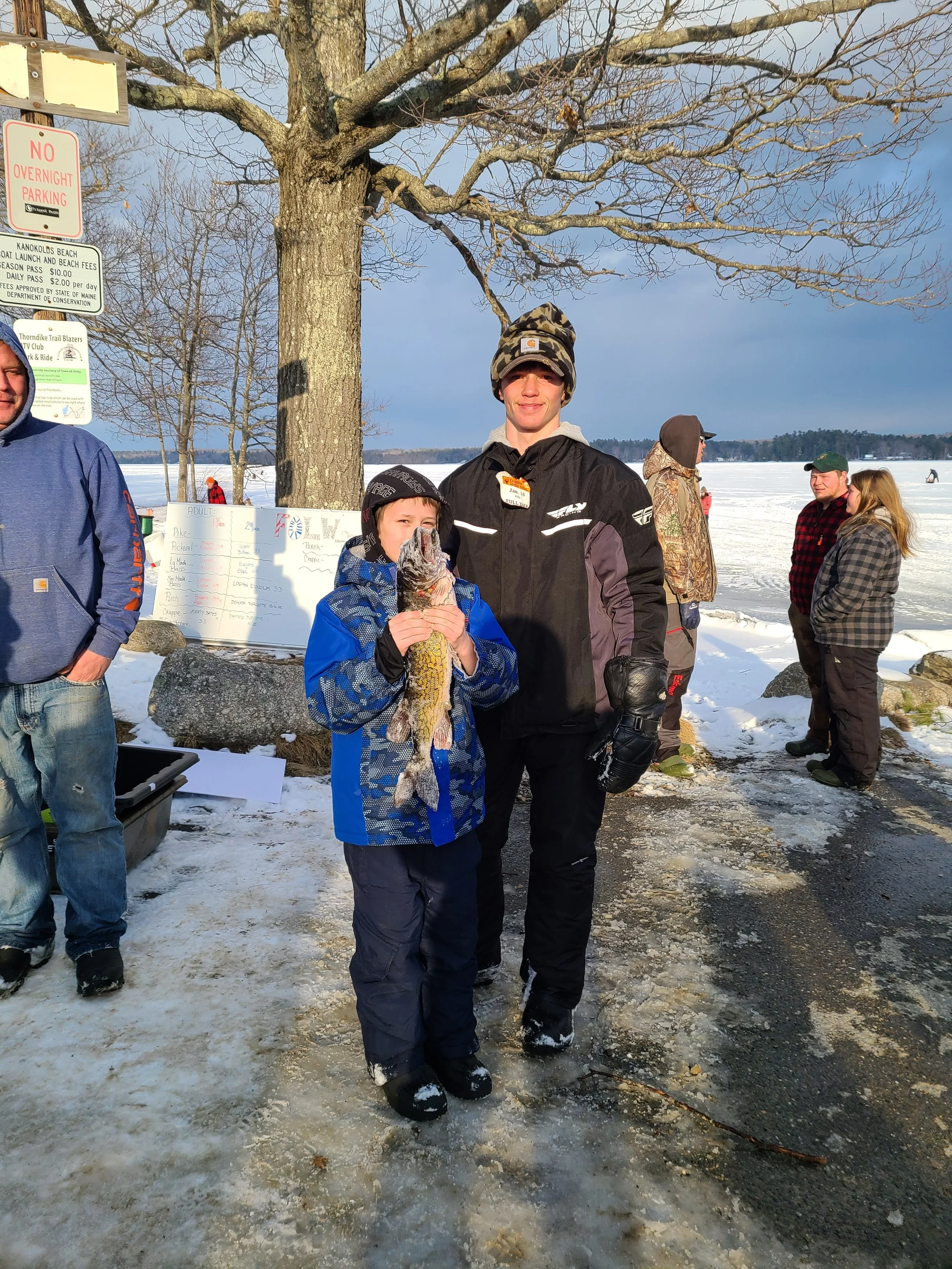 A young boy holding a freshly caught fish, standing next to a smiling young man with winter clothing outdoors on a snowy day near a frozen lake, with other people in the background.