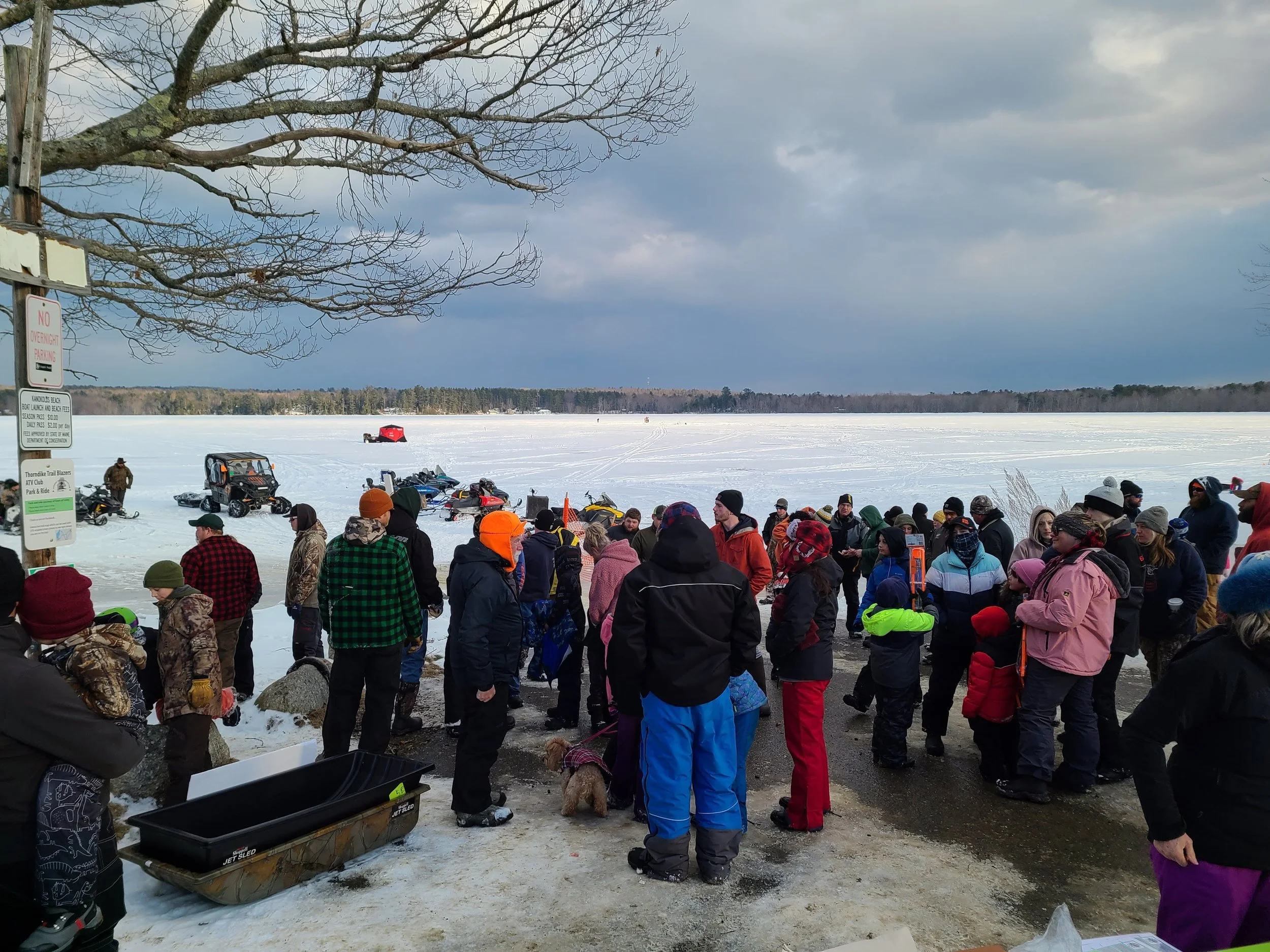 Group of people gathered near a frozen lake with snowmobiles in the background, overcast sky, winter clothing, some children and adults, with trees and a snow-covered landscape.