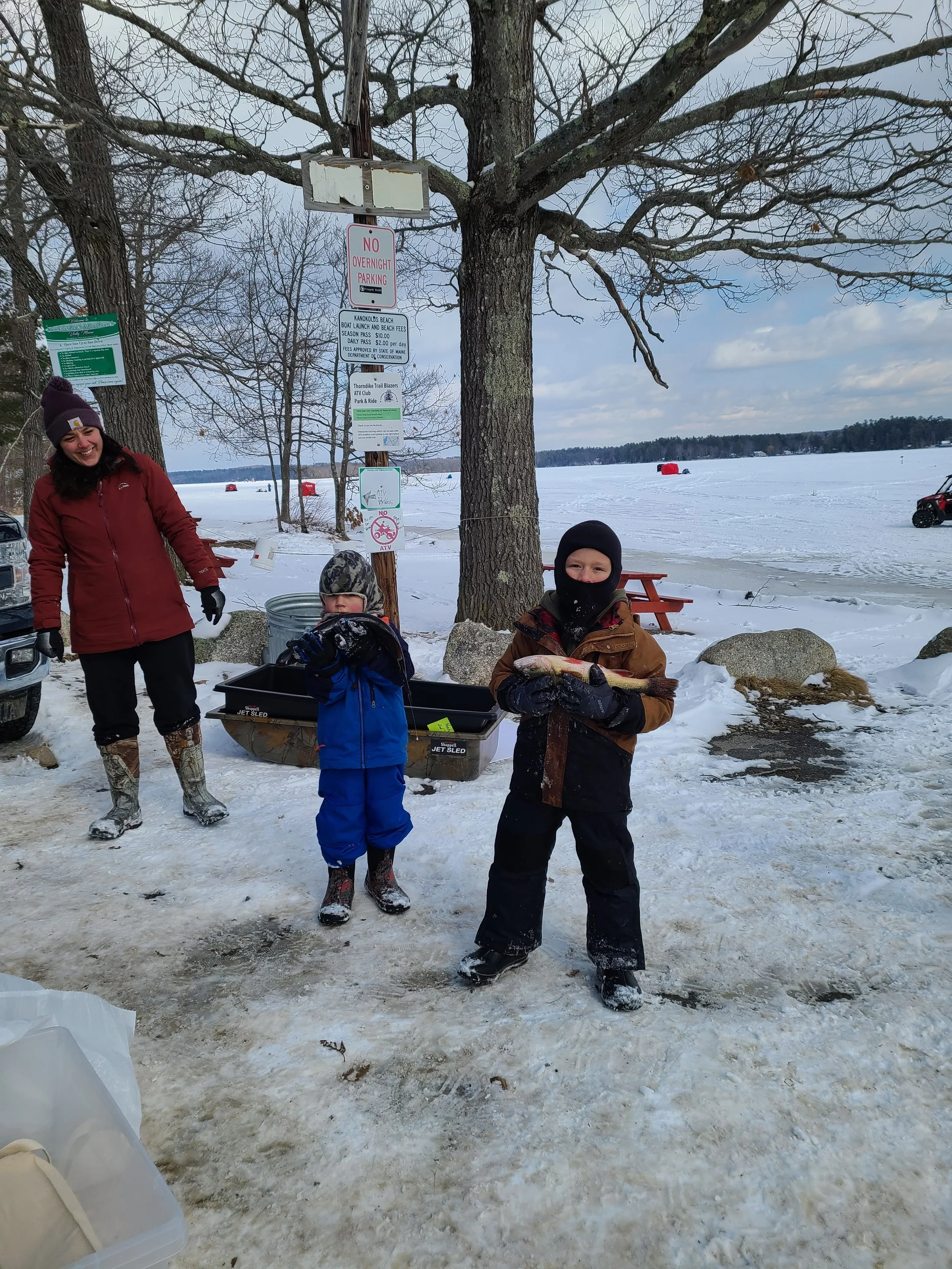Two children and an adult in winter clothing standing on snow-covered ground near a frozen lake, with the older child holding a fish and the younger child standing beside him. An adult woman in winter gear is smiling nearby, and there are trees and s