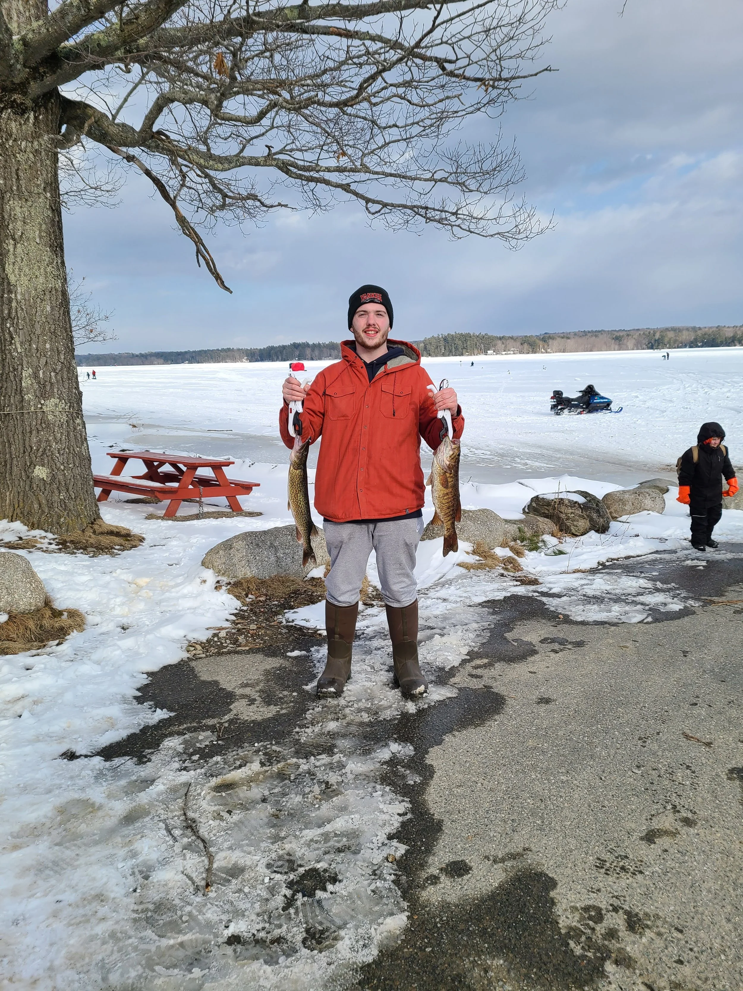 A young man wearing a black beanie, red jacket, gray sweatpants, and brown boots stands outdoors on snow and ice, holding two large fish, one in each hand. There is a large tree, a red picnic table, and people in the background on a snowy, frozen lak