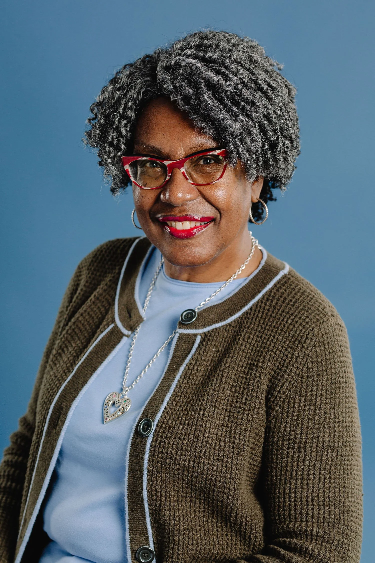 A woman with gray, curly hair wearing red glasses, a brown cardigan, a light blue shirt, and heart-shaped jewelry, smiling against a blue background.