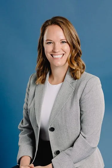 A woman with shoulder-length brown hair smiling, wearing a gray blazer and white shirt against a blue background.