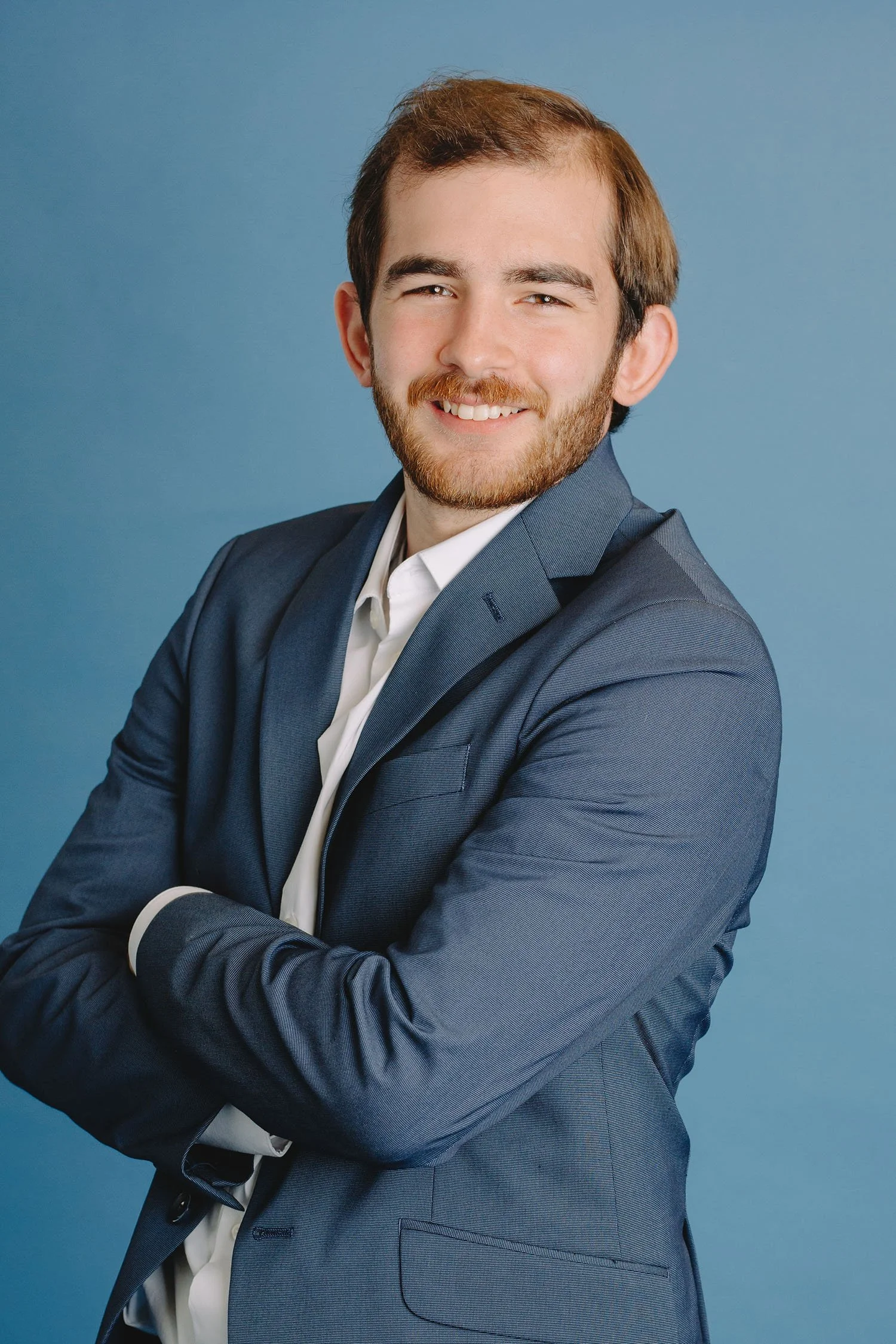 A young man with a beard and short brown hair, wearing a blue suit and white shirt, smiling with arms crossed, standing against a blue background.