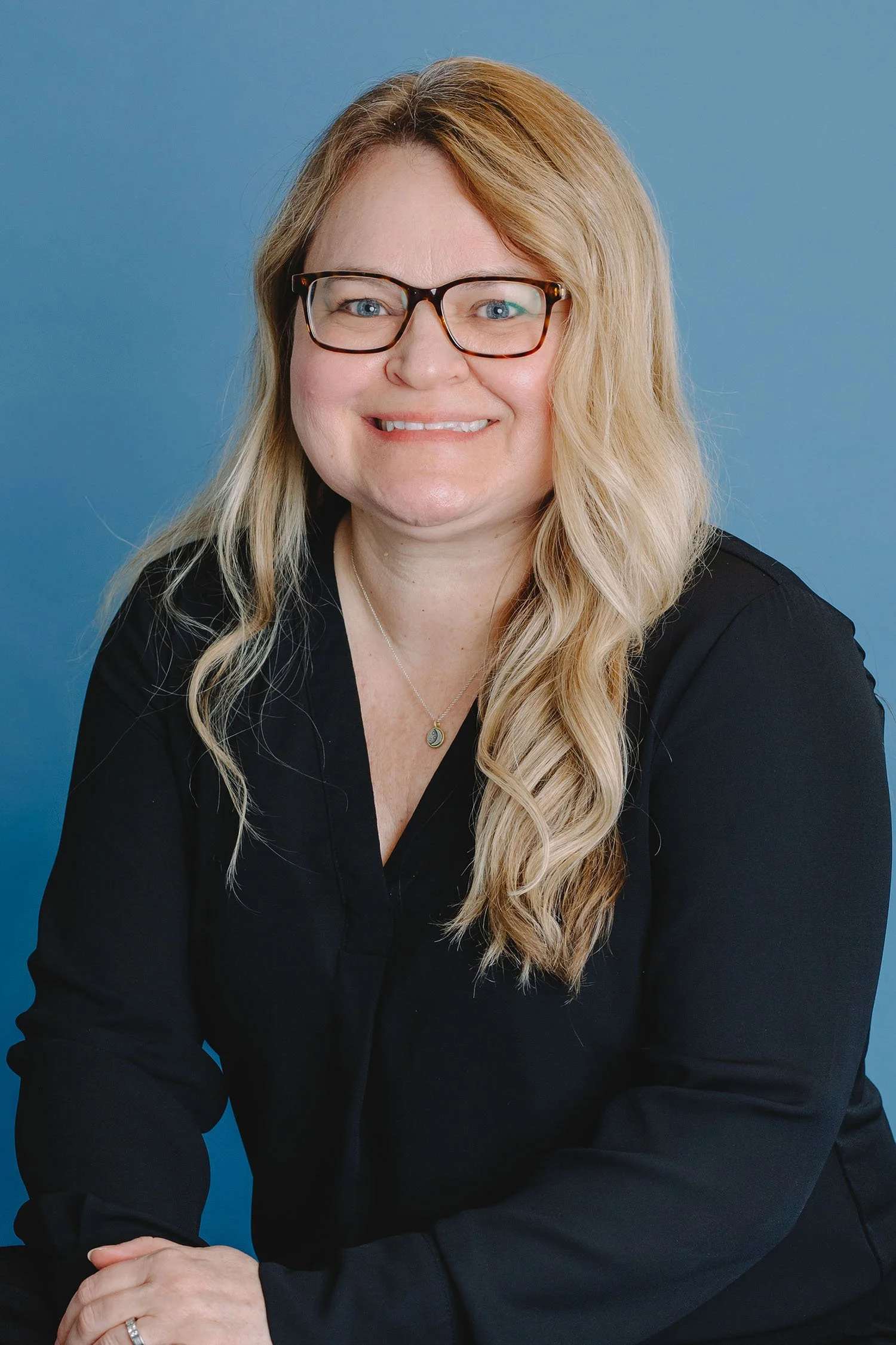 Portrait of a woman with long blonde hair, wearing glasses, a black shirt, and a necklace, smiling against a blue background.
