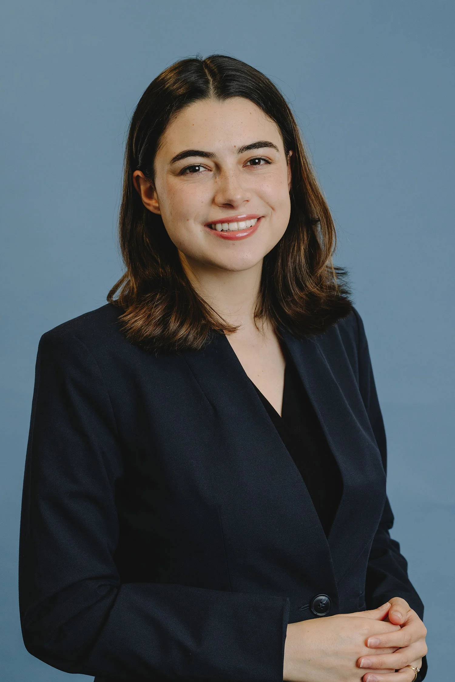 A woman with shoulder-length brown hair, wearing a dark blazer, smiling in front of a plain blue background.