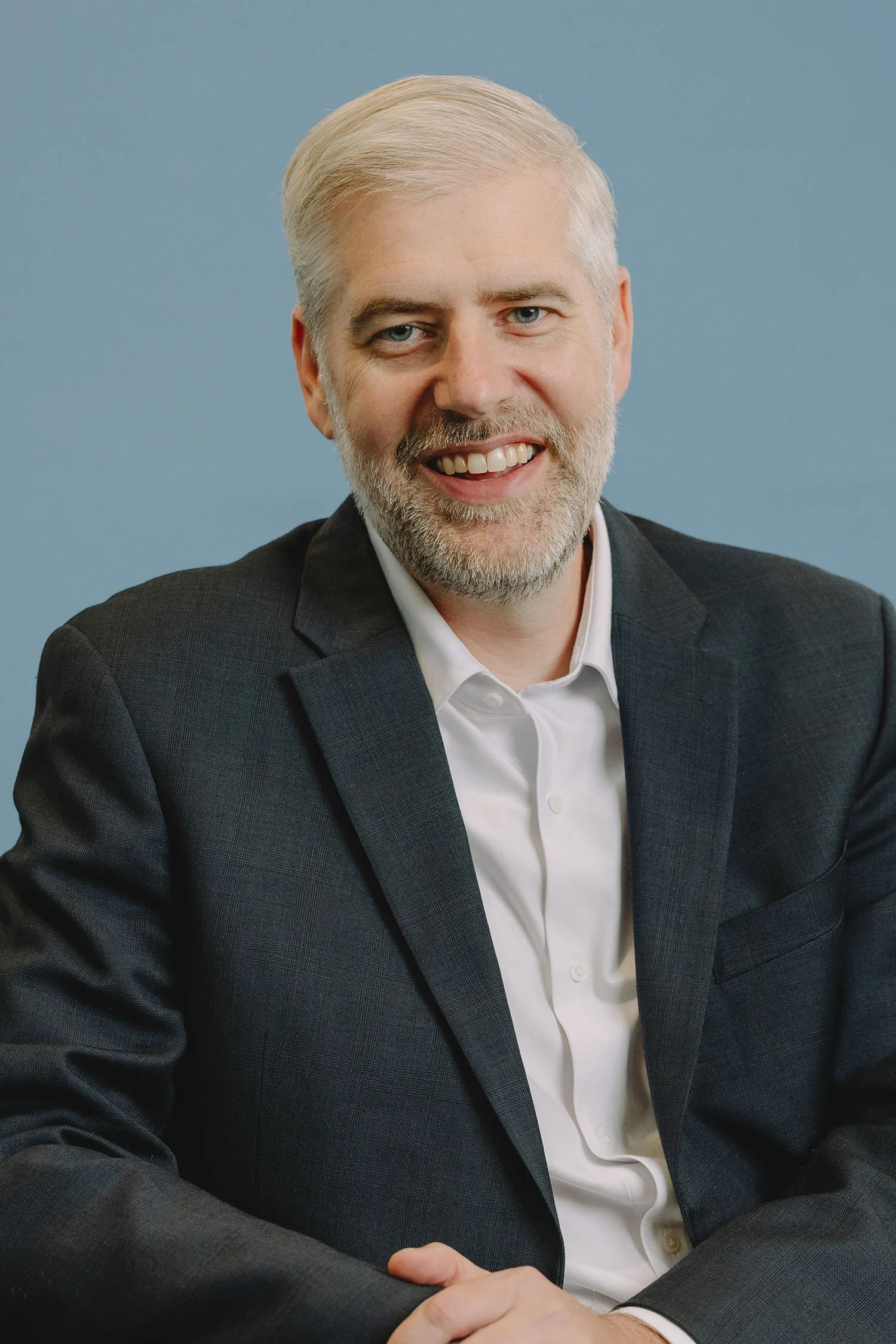 A middle-aged man with gray hair and a beard smiling, wearing a dark suit jacket and white shirt, sitting against a blue background.