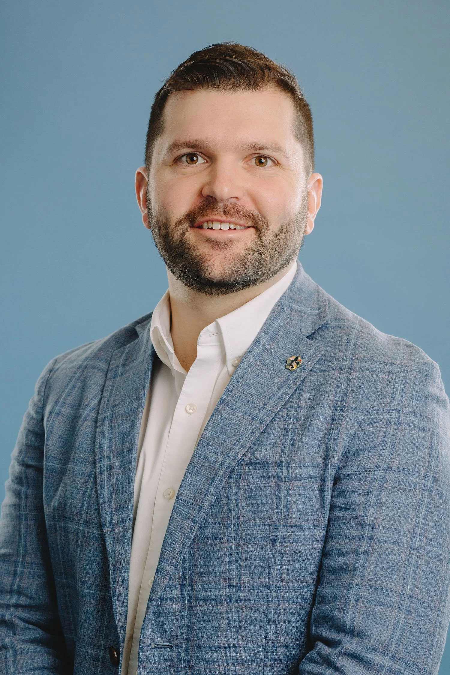 Portrait of a man with short dark hair, beard, and brown eyes, wearing a light gray blazer with a small lapel pin and a white collared shirt against a blue background.